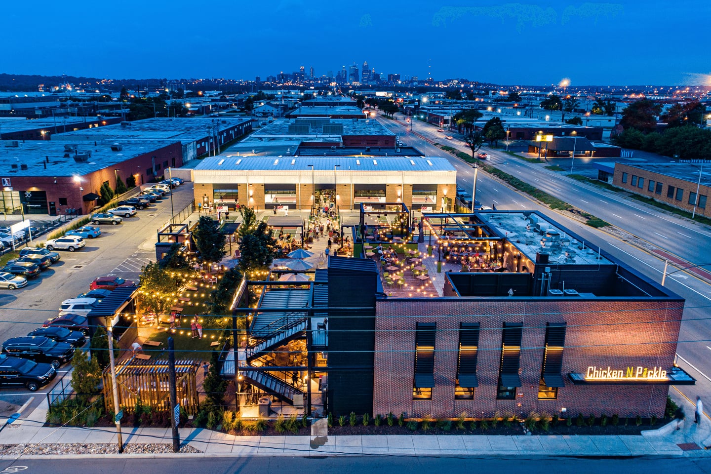 Aerial view of a commercial building with outdoor seating and string lights, parking lot, and surrounding industrial area at dusk.