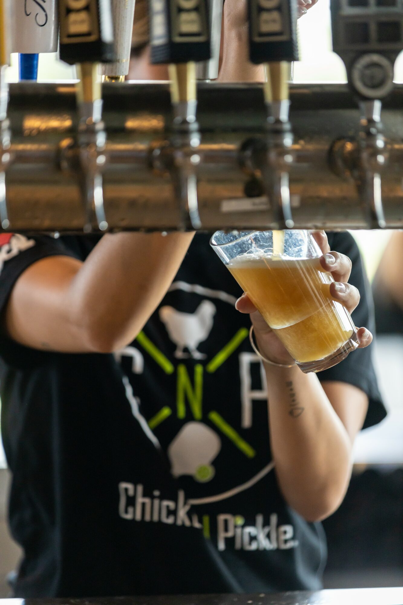Person holding a glass of beer at a bar with beer taps above, wearing a black T-shirt with a chicken and pickle design.