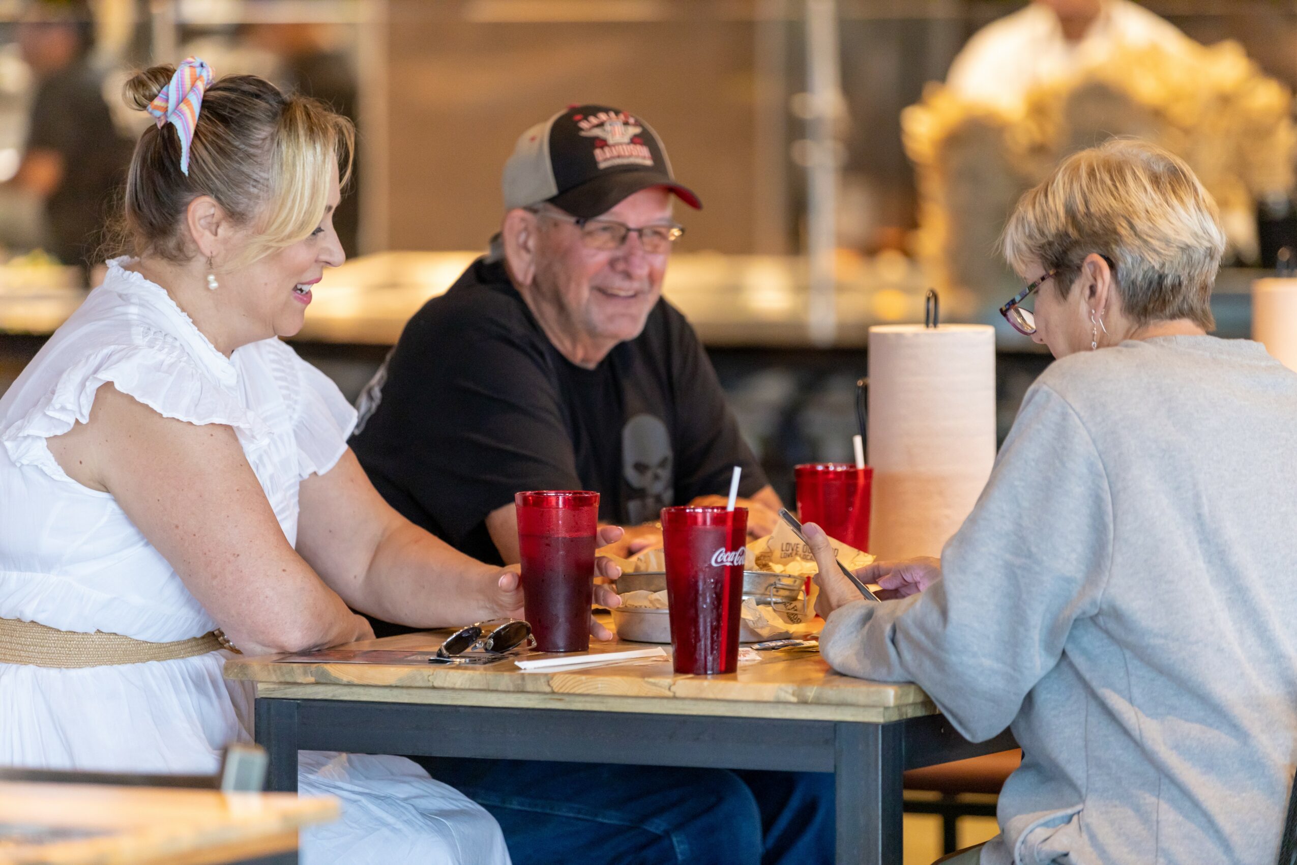 Three people sitting at a table in a restaurant, smiling and talking, with drinks and a napkin holder on the table.