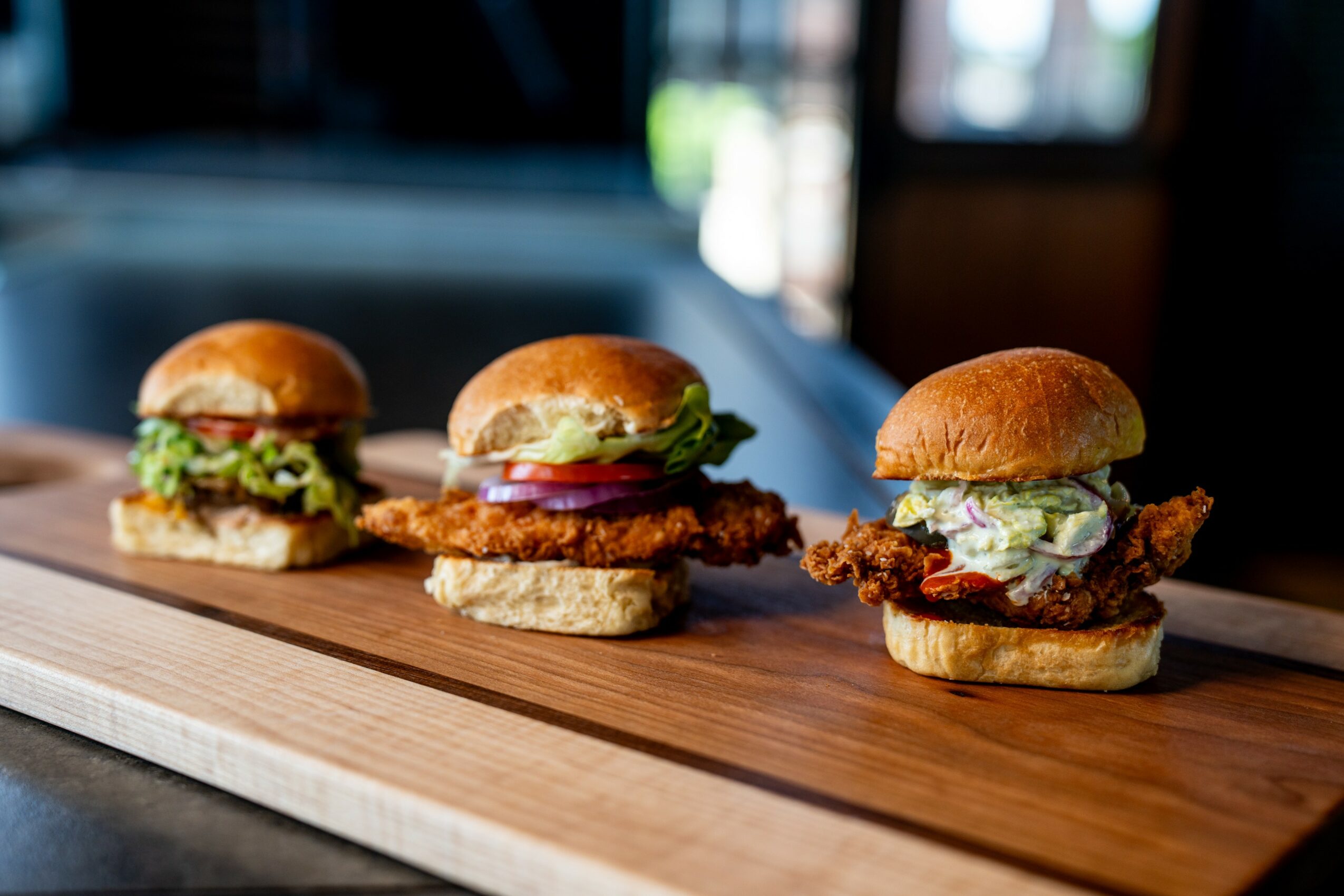 Three sandwiches on a wooden board, each with different fillings and buns, in a cozy indoor setting.