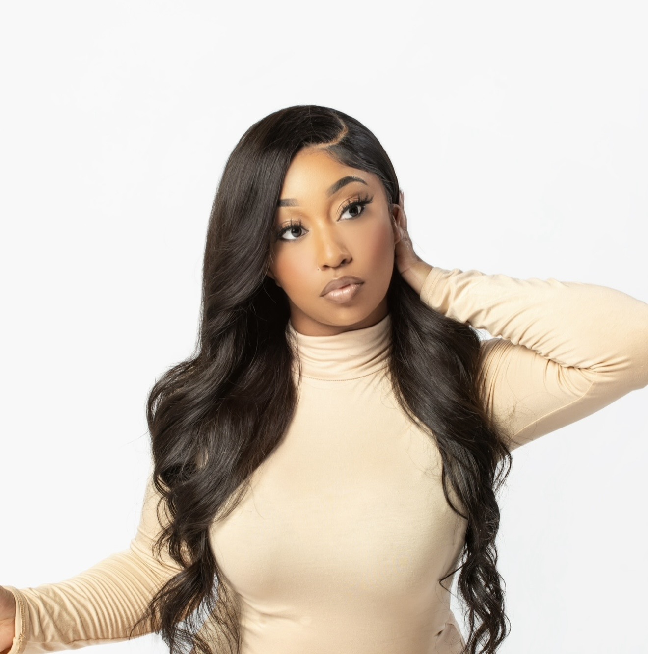 Woman with long wavy hair wearing a beige turtleneck, looking to the side, hand near her neck, plain white background.