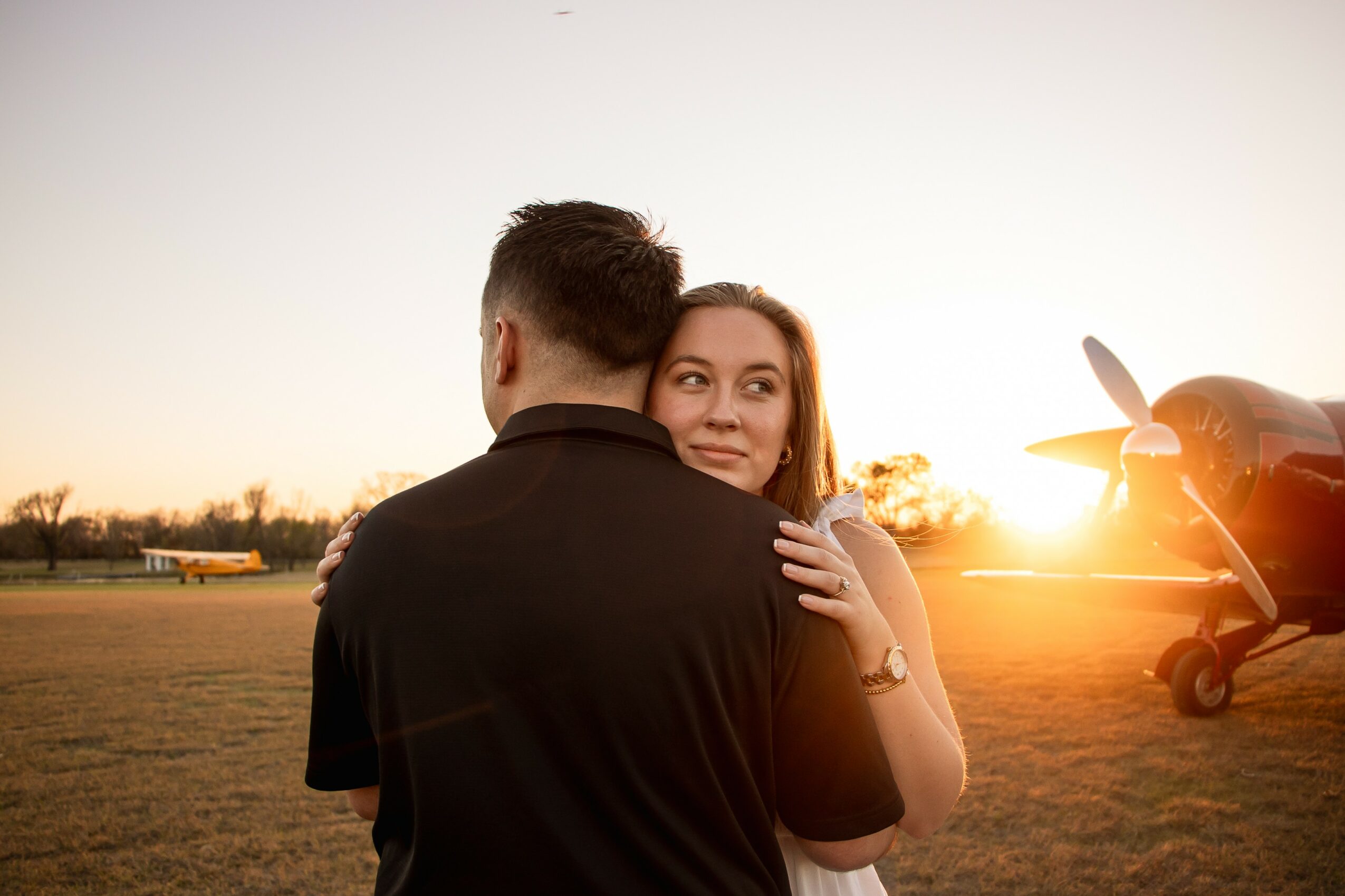 A woman embraces a man outdoors during sunset, with an airplane and open field in the background.