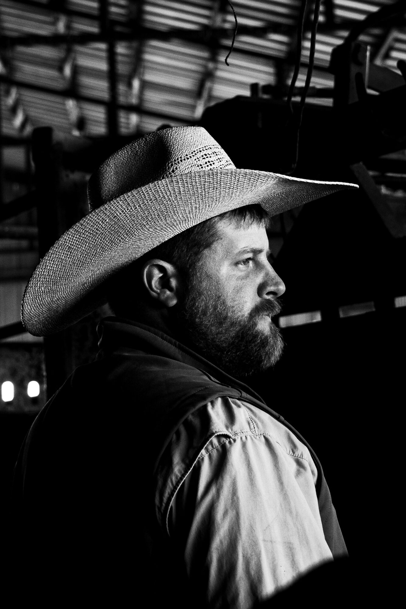 Man with a beard wearing a wide-brimmed hat, looking to the right, in a dark setting with structural elements overhead.