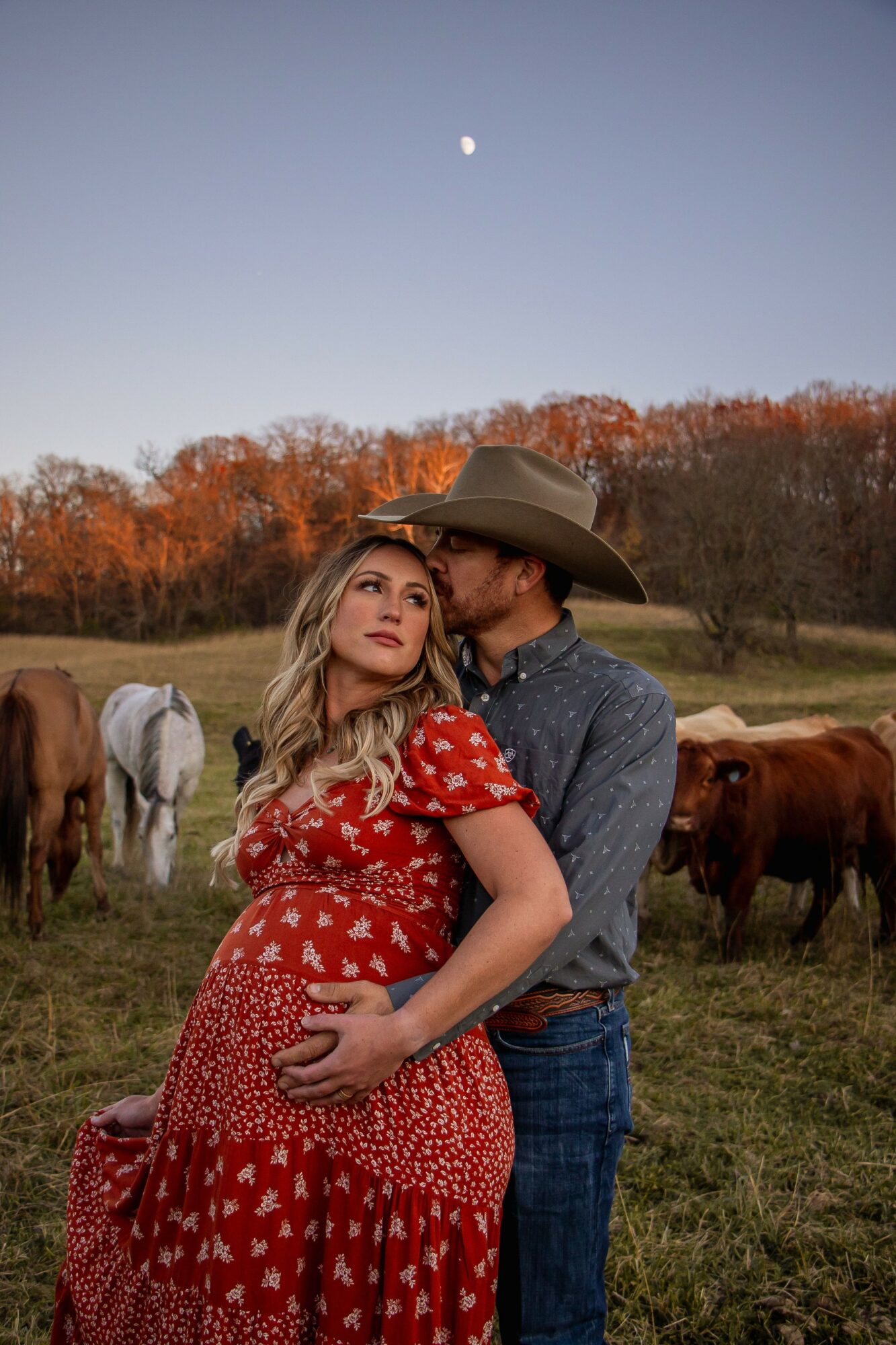 A couple stands in a field with horses, the man wearing a cowboy hat and the woman in a red dress, during sunset.