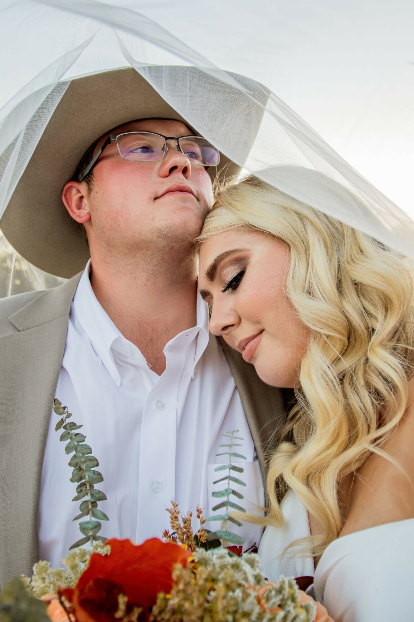 A couple with closed eyes under a veil, holding a bouquet, outdoors, with soft lighting.