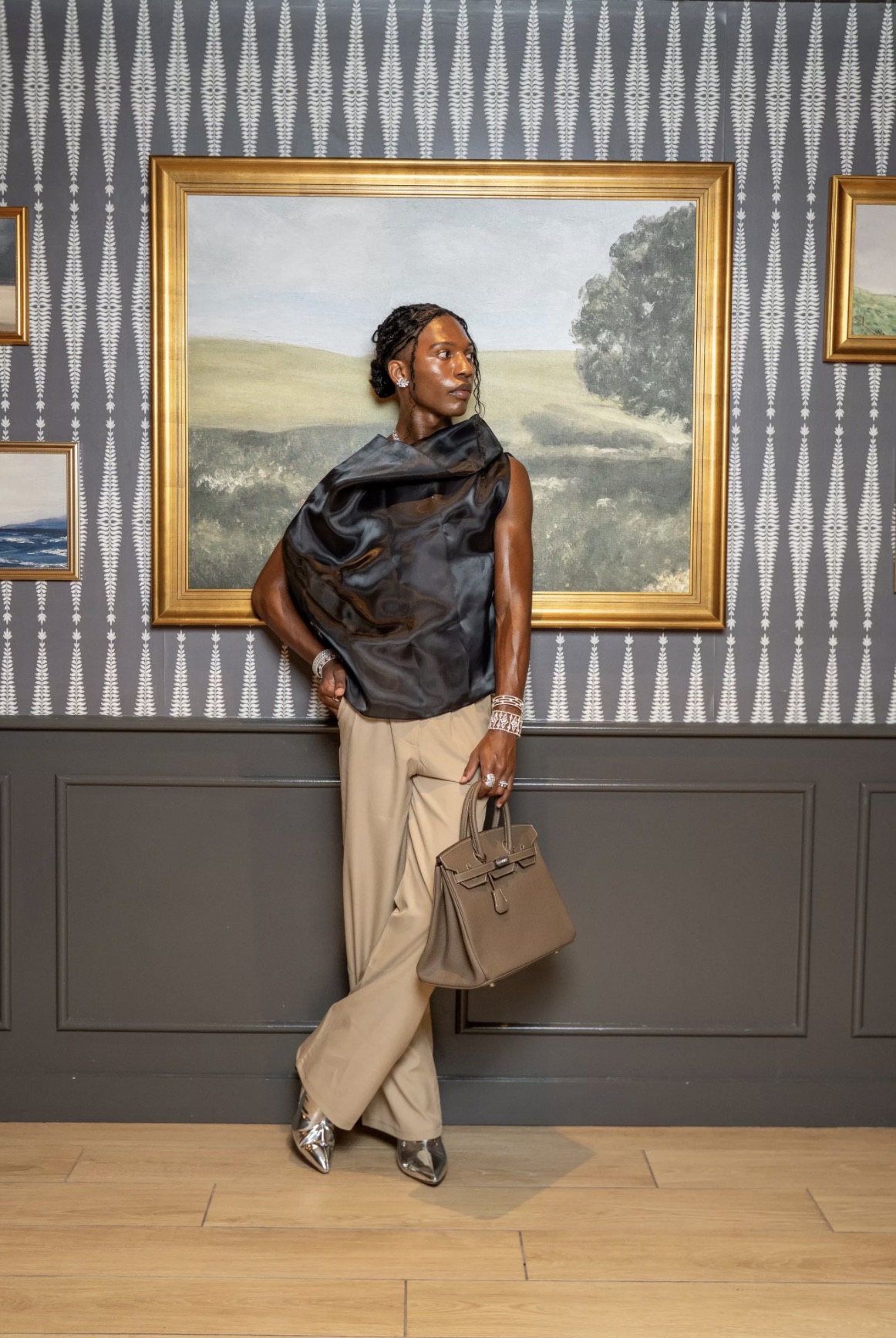 Woman standing in art gallery, looking at landscape painting, holding a handbag, wearing beige pants and black top.