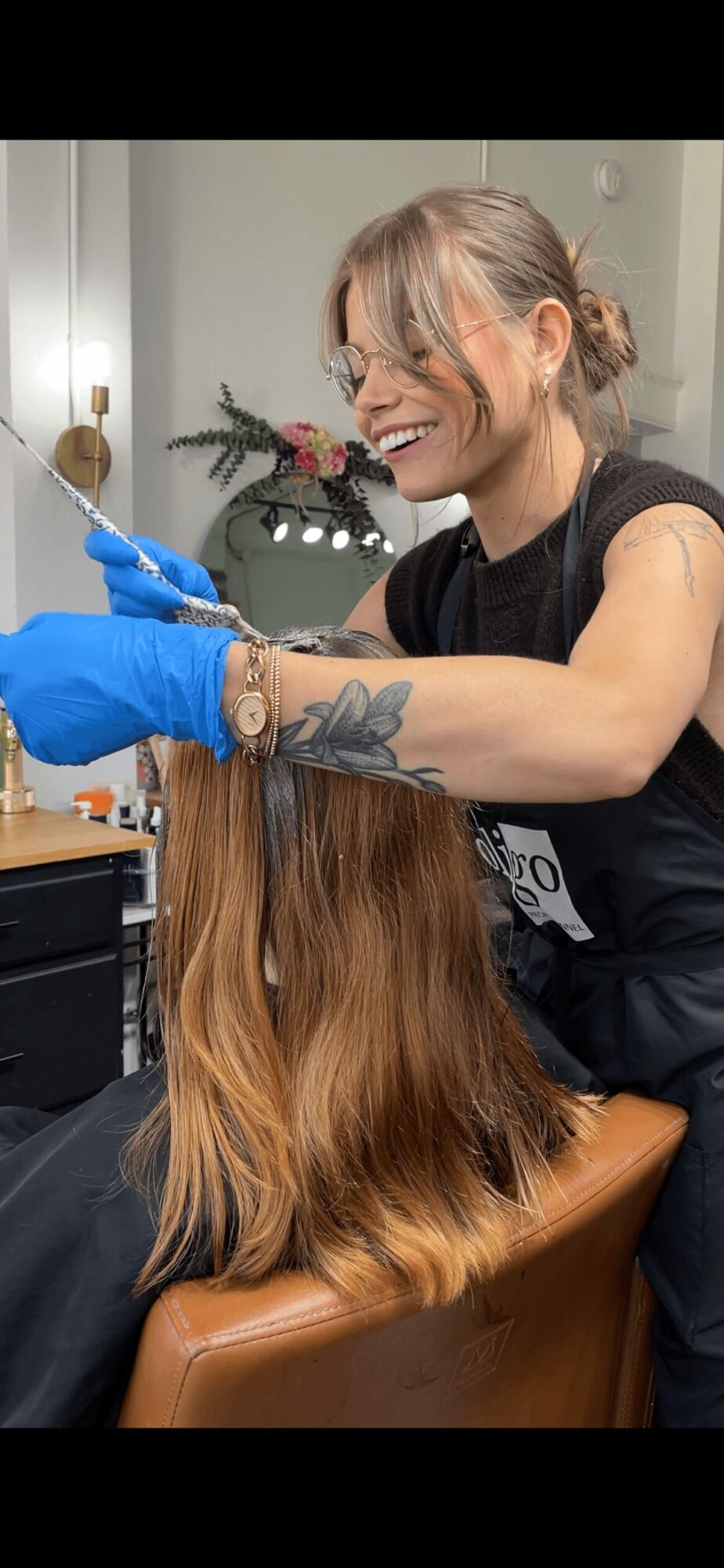 Woman with glasses and a tattoo on her arm smiling while working on a client's hair, wearing blue gloves.