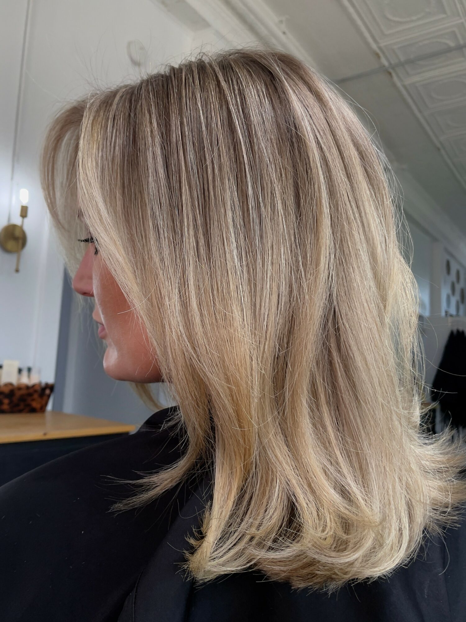 Side view of a woman with shoulder-length blonde hair styled in loose waves, indoors with a white ceiling and wall.