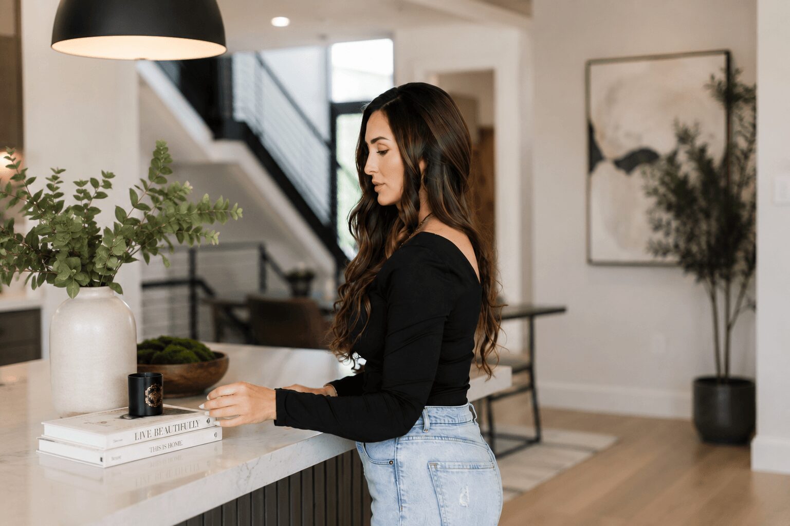 Woman with long dark hair in black top and jeans stands at a kitchen counter, holding a small object.