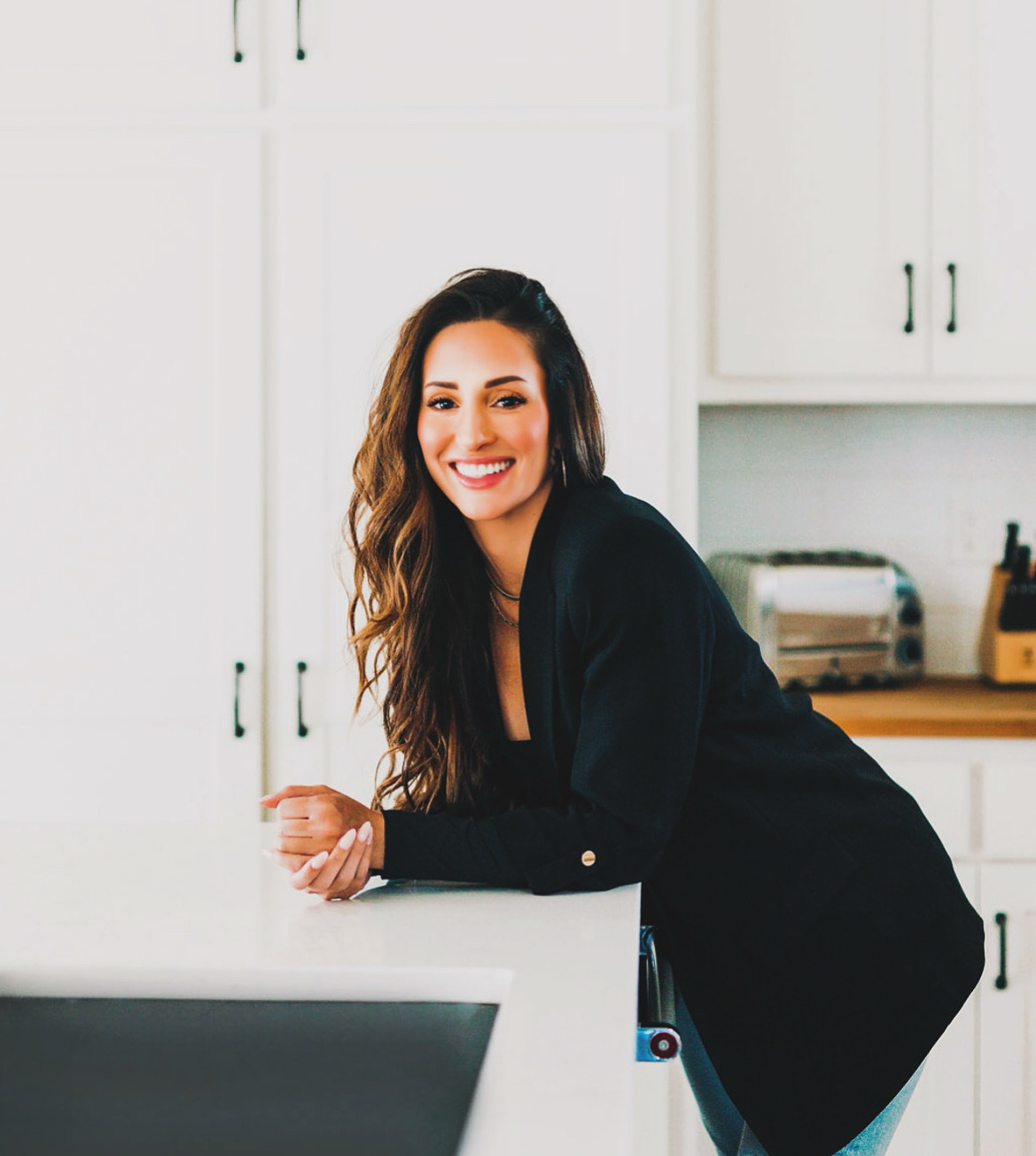 Smiling woman with long hair leaning on a kitchen counter in a bright, white kitchen with cabinets and appliances.