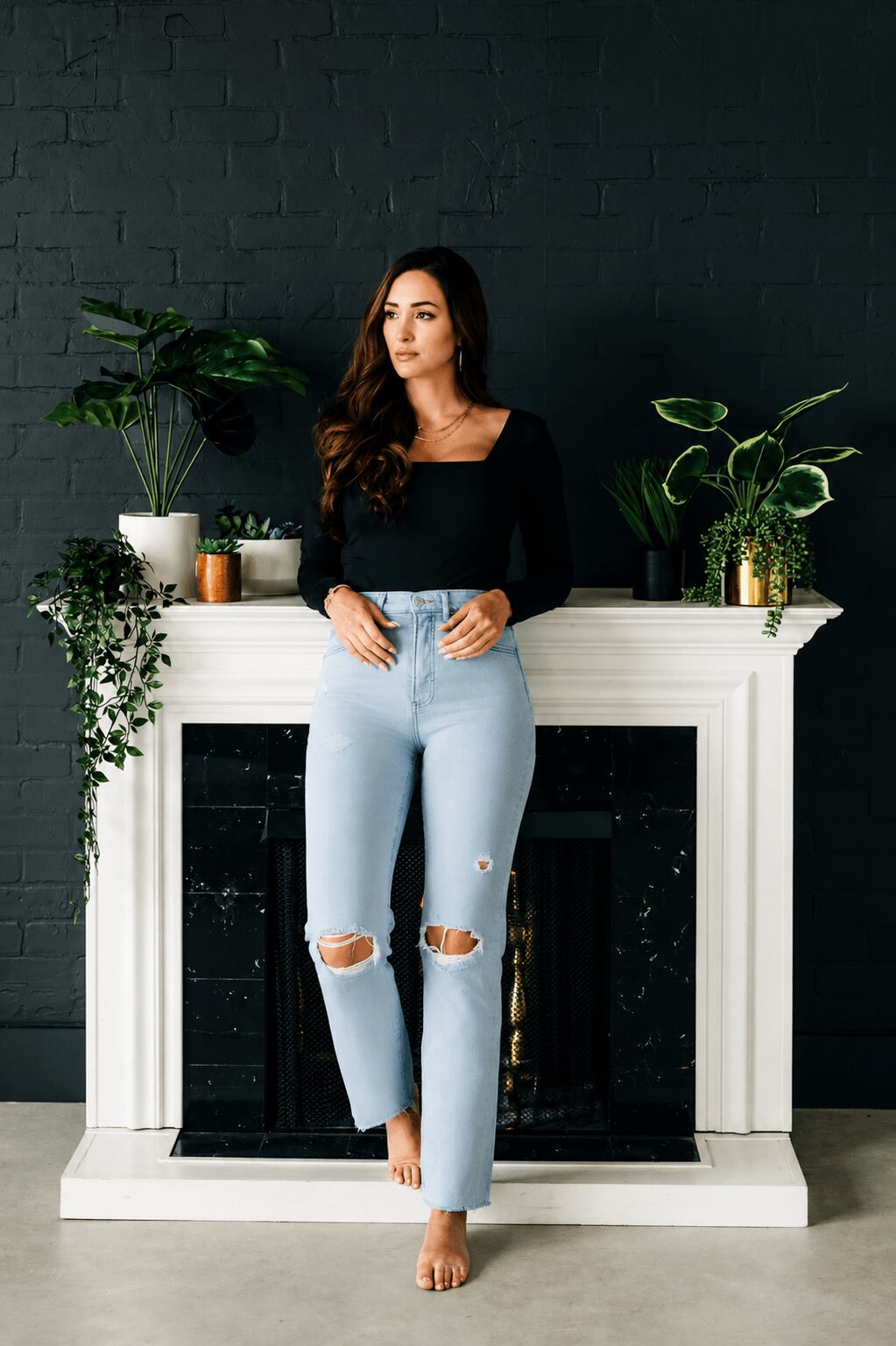 Woman with long dark hair in black top and ripped jeans stands in front of a white fireplace with plants, against a dark wall.