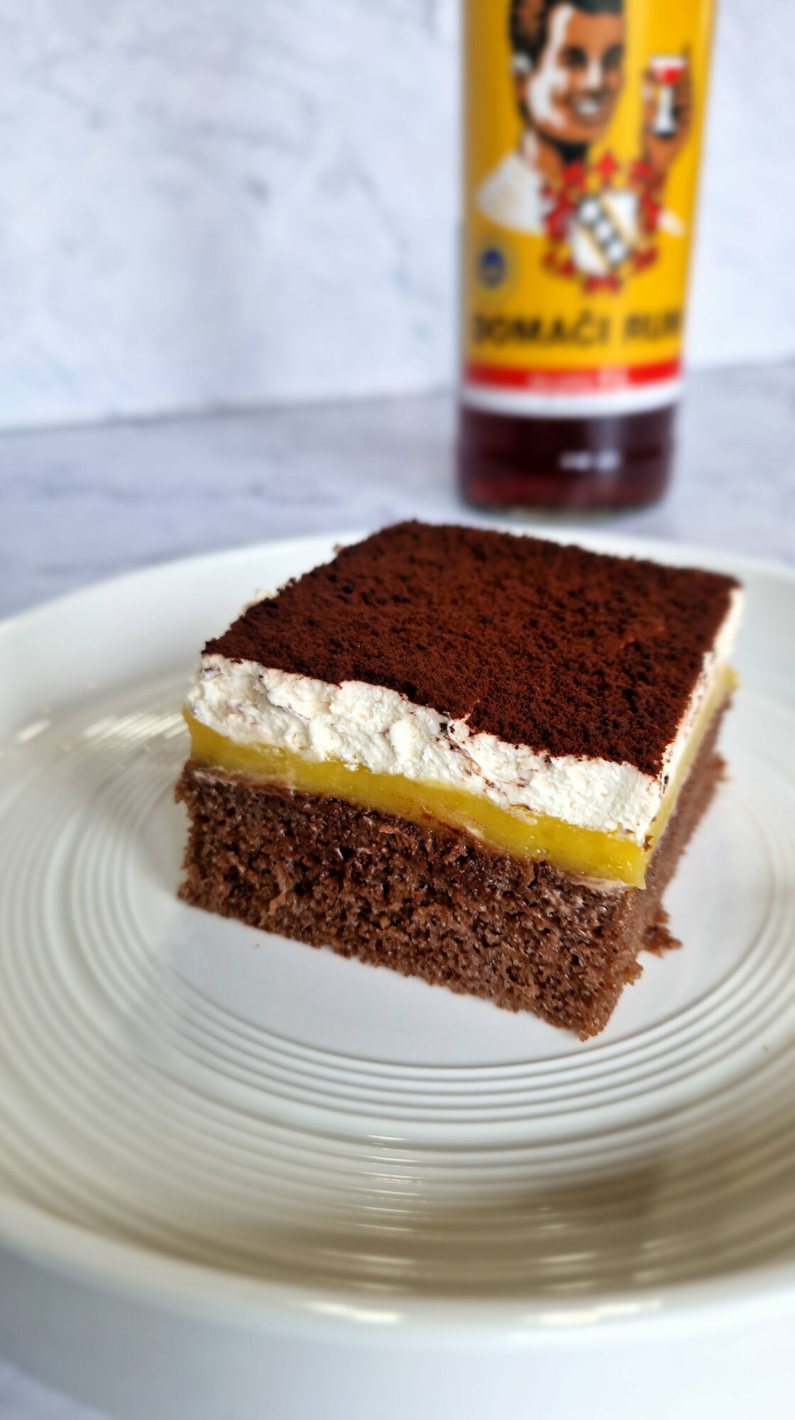 A slice of chocolate cake with cream and lemon filling on a clear plate, with a bottle of hot sauce nearby.