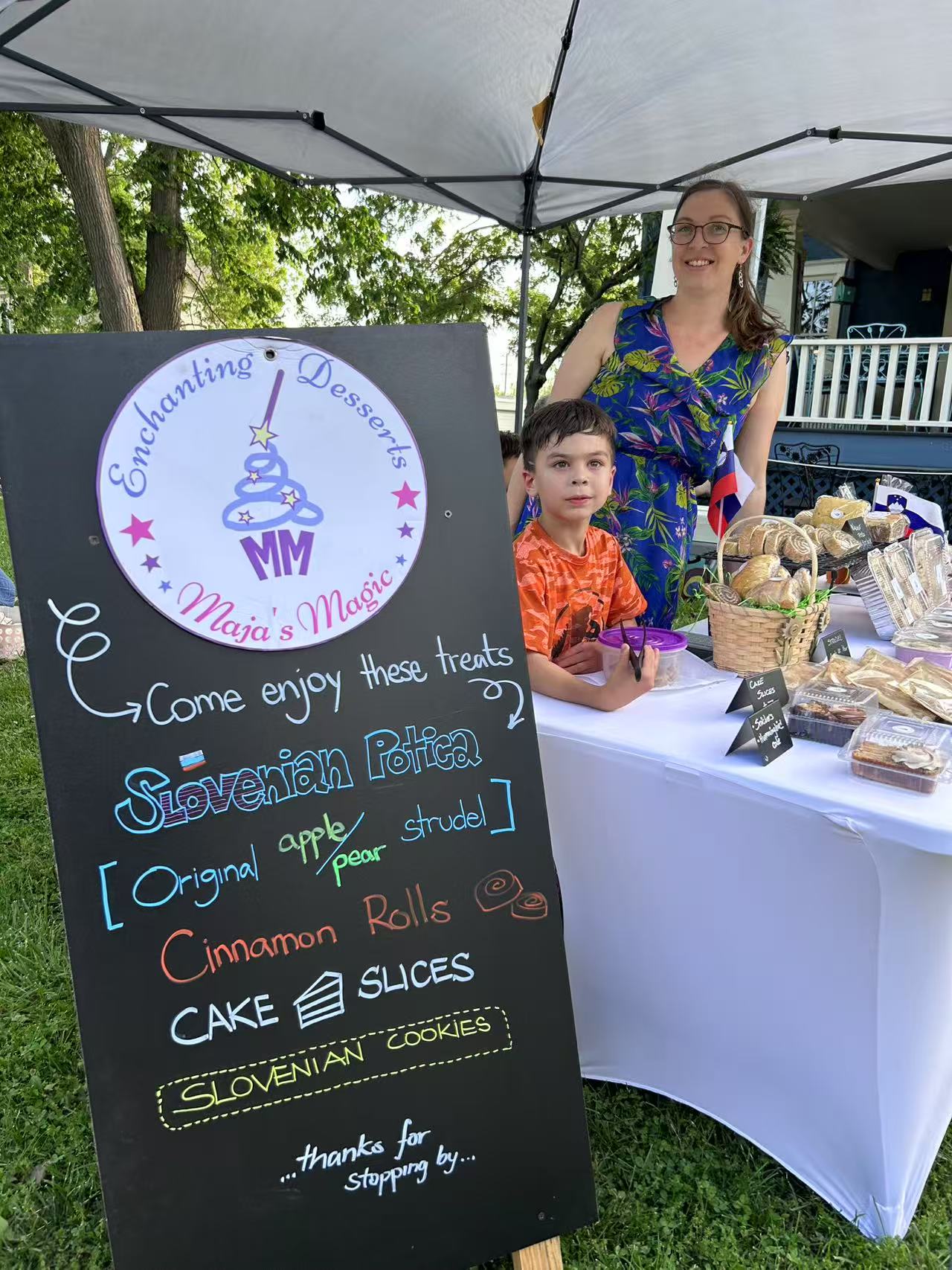 Woman and child at a table with a blackboard sign outdoors, surrounded by trees and a white tent, smiling.