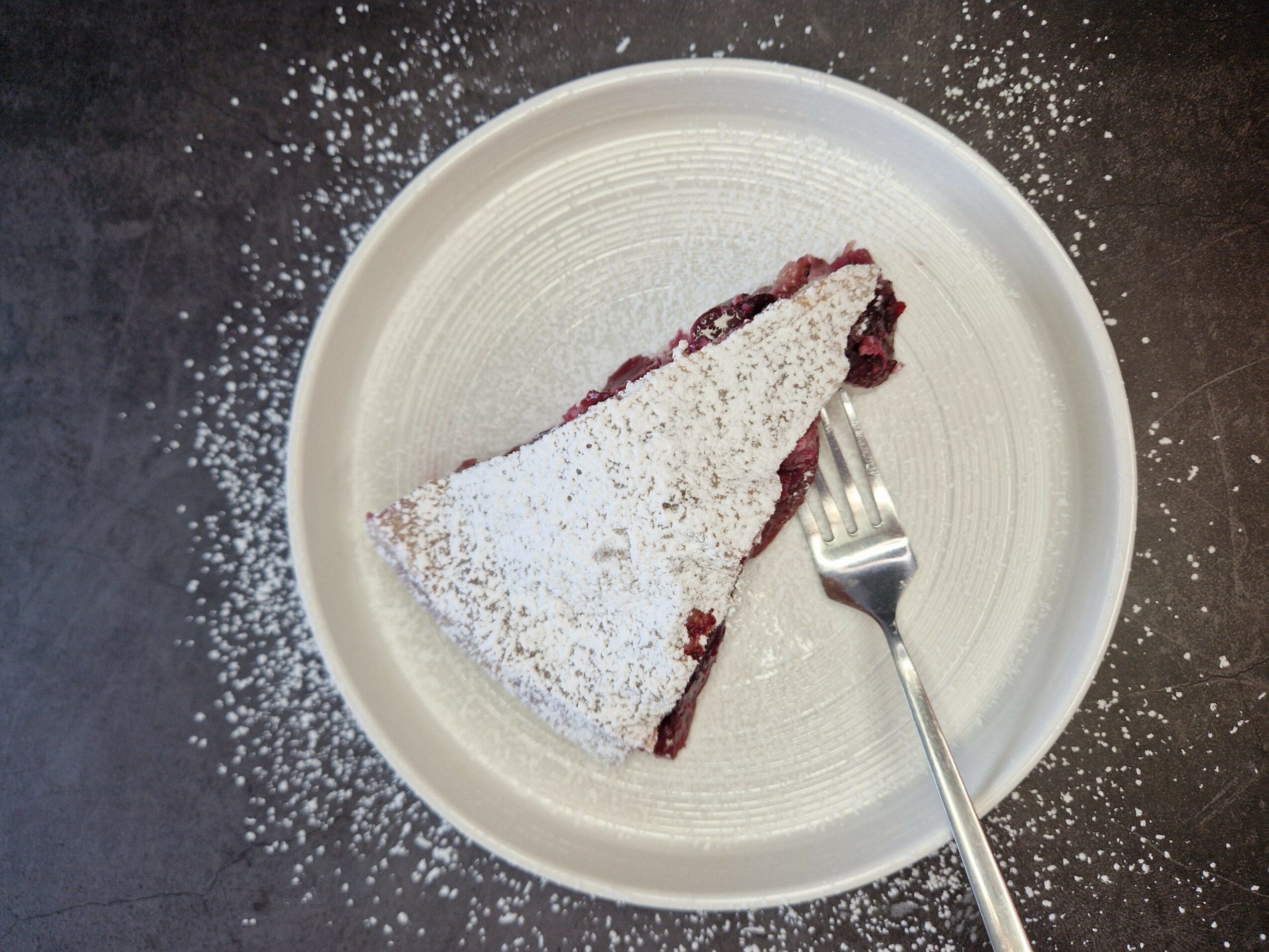 A slice of cherry pie with powdered sugar on a white plate, with scattered sugar on dark surface, fork on plate.