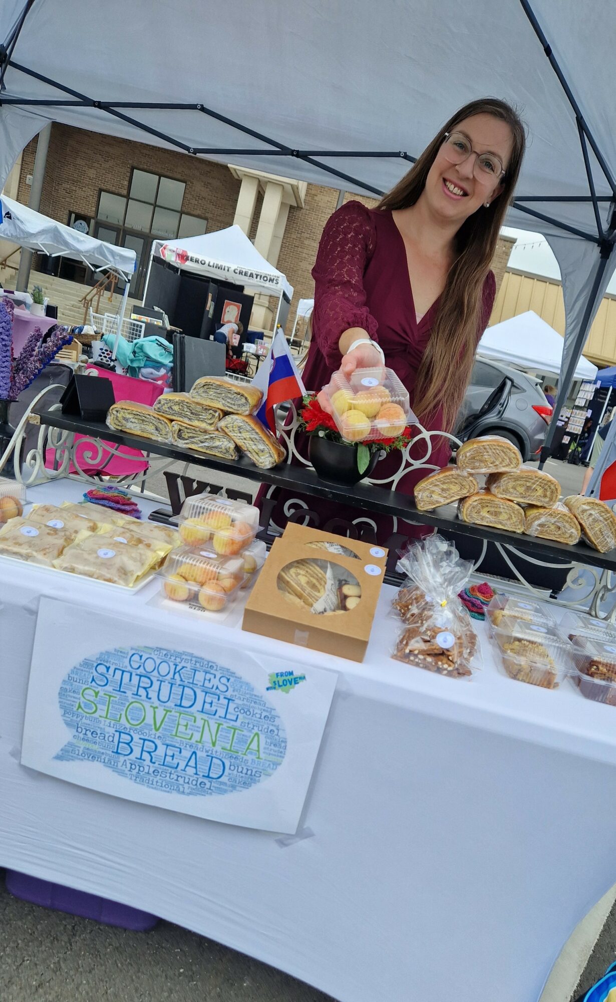 Woman smiling behind a table with baked goods and colorful candies at an outdoor market.
