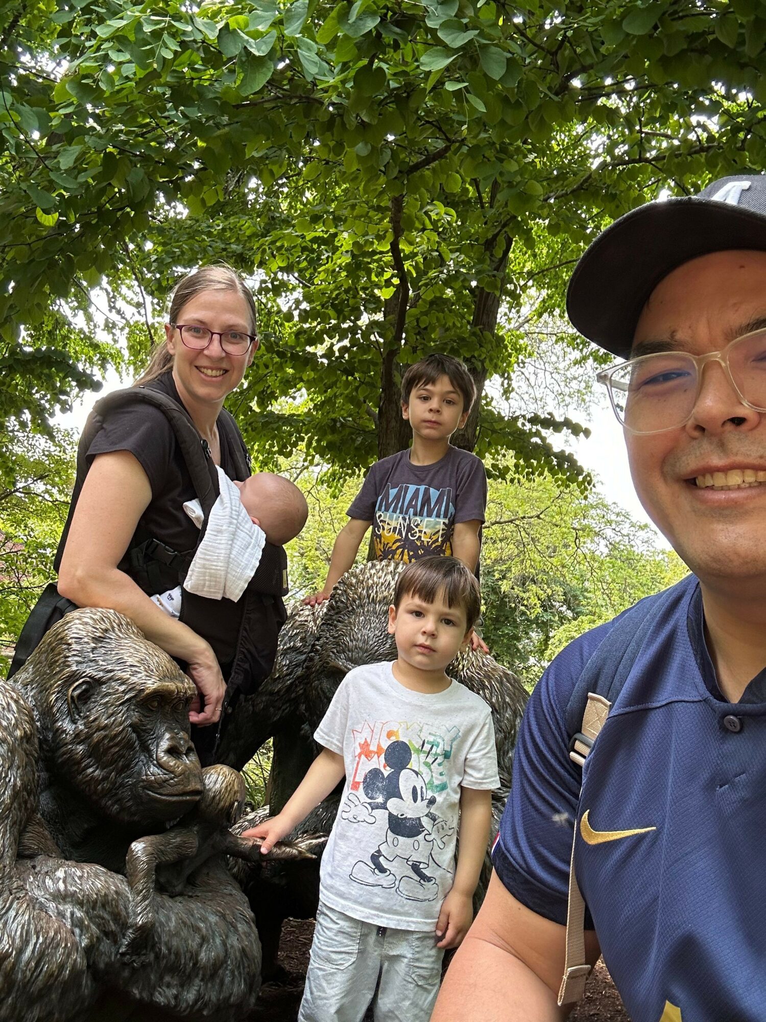 Group of children and a woman outdoors with a dog, surrounded by green trees, smiling and looking at the camera.