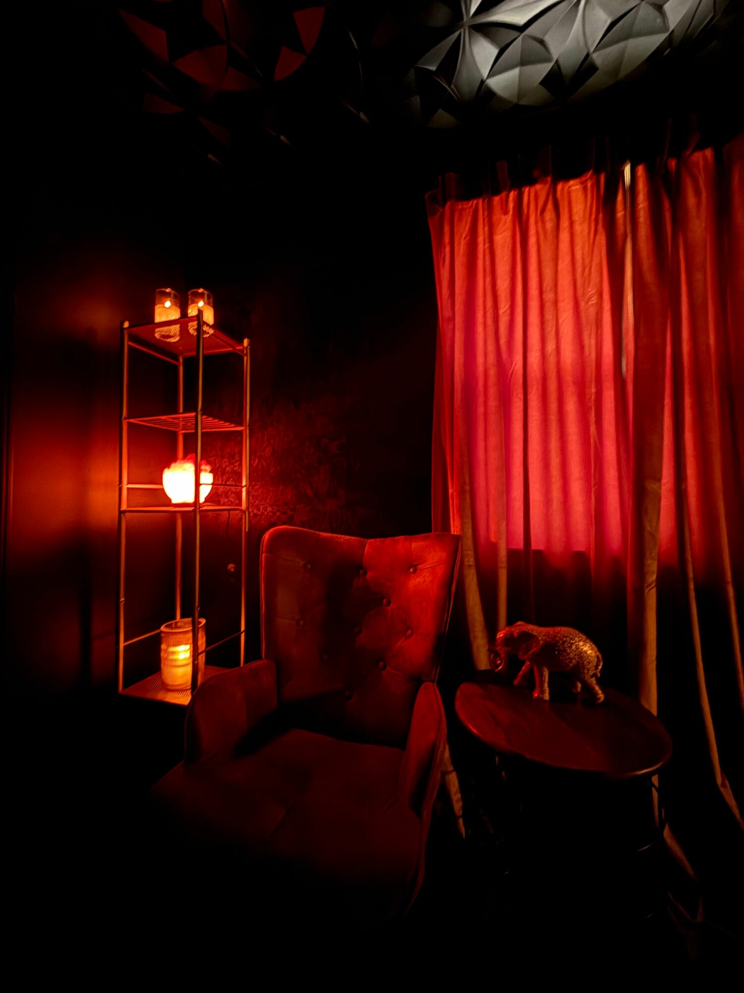 Dimly lit room with red curtains, a table with a teddy bear, and a glowing light fixture on the wall.