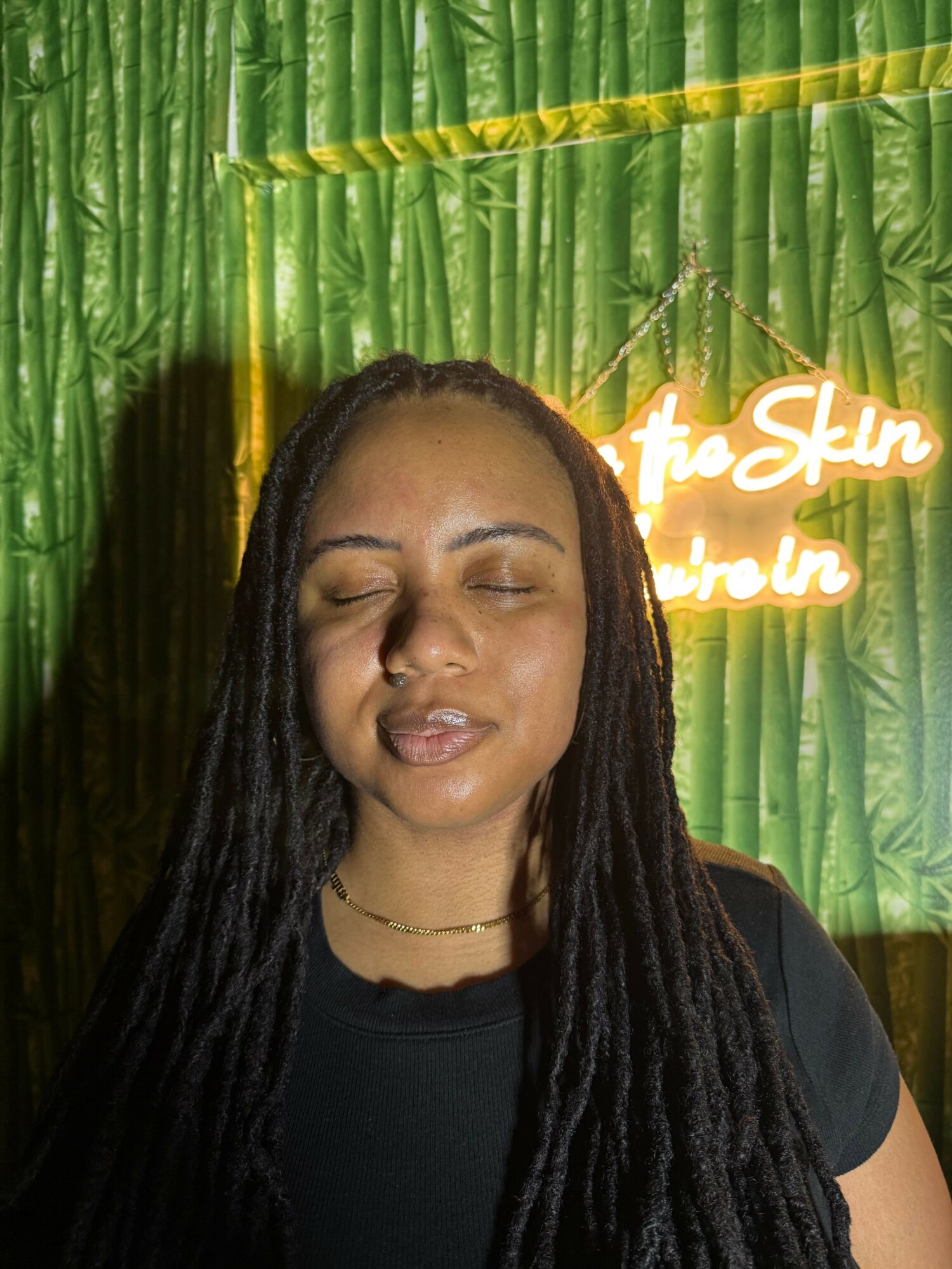 Woman with long dreadlocks and closed eyes, standing in front of a green textured background with neon sign, lips puckered.