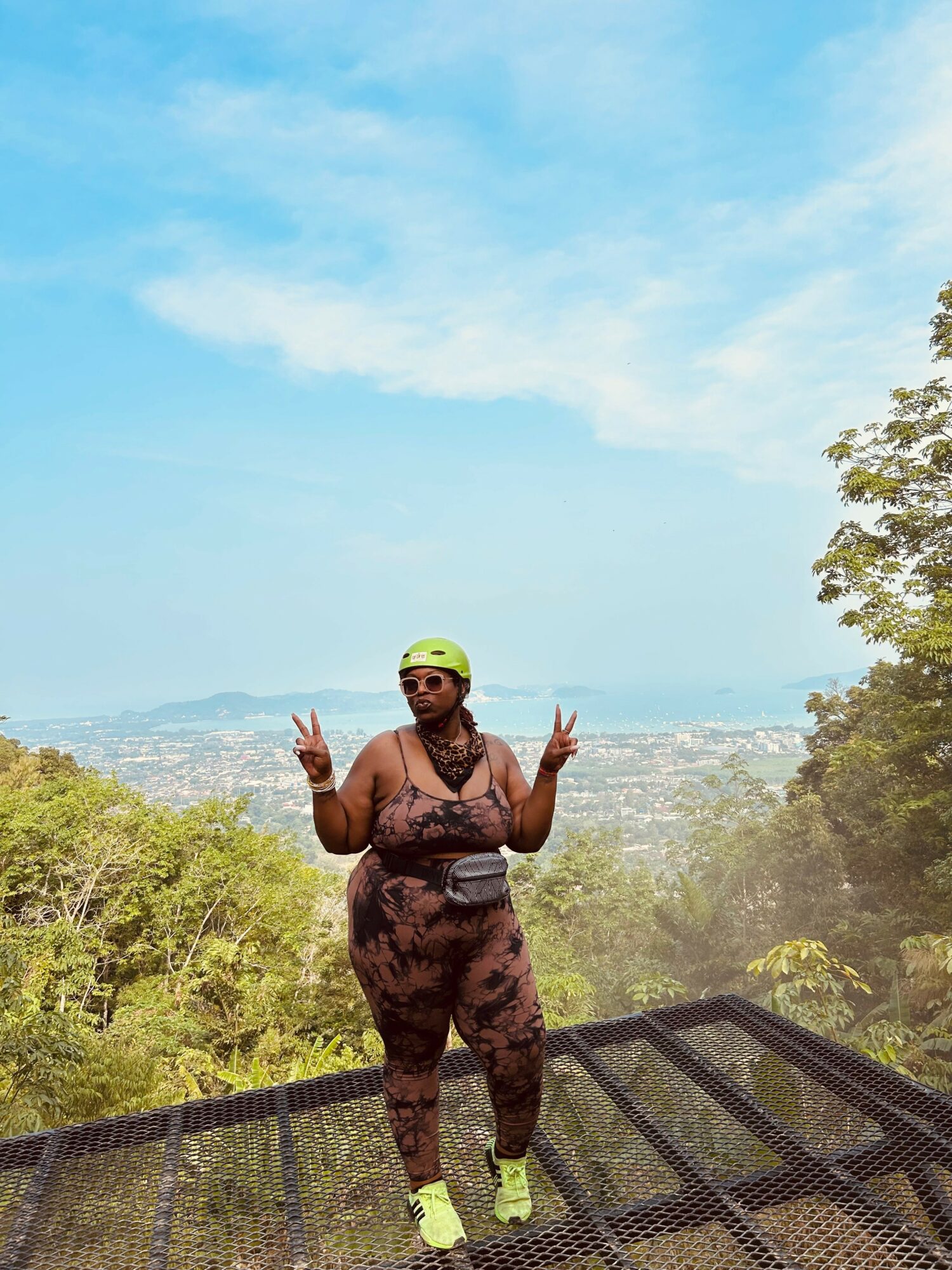 Person standing on a metal platform outdoors, making peace signs with both hands, wearing sunglasses, a helmet, and athletic clothing, with trees and sky in background.