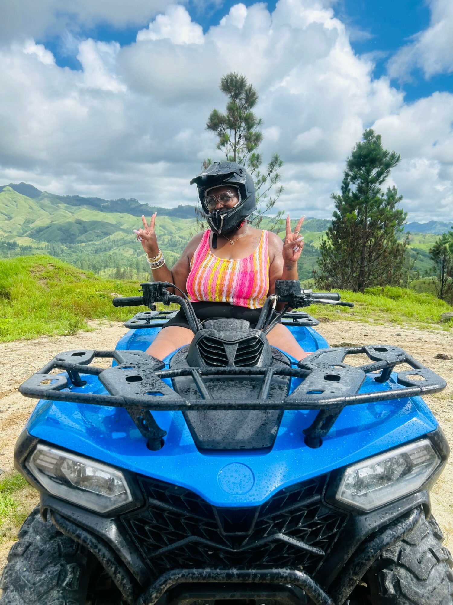 Person wearing a helmet and pink striped top sitting on a blue ATV outdoors with green hills and trees in the background.