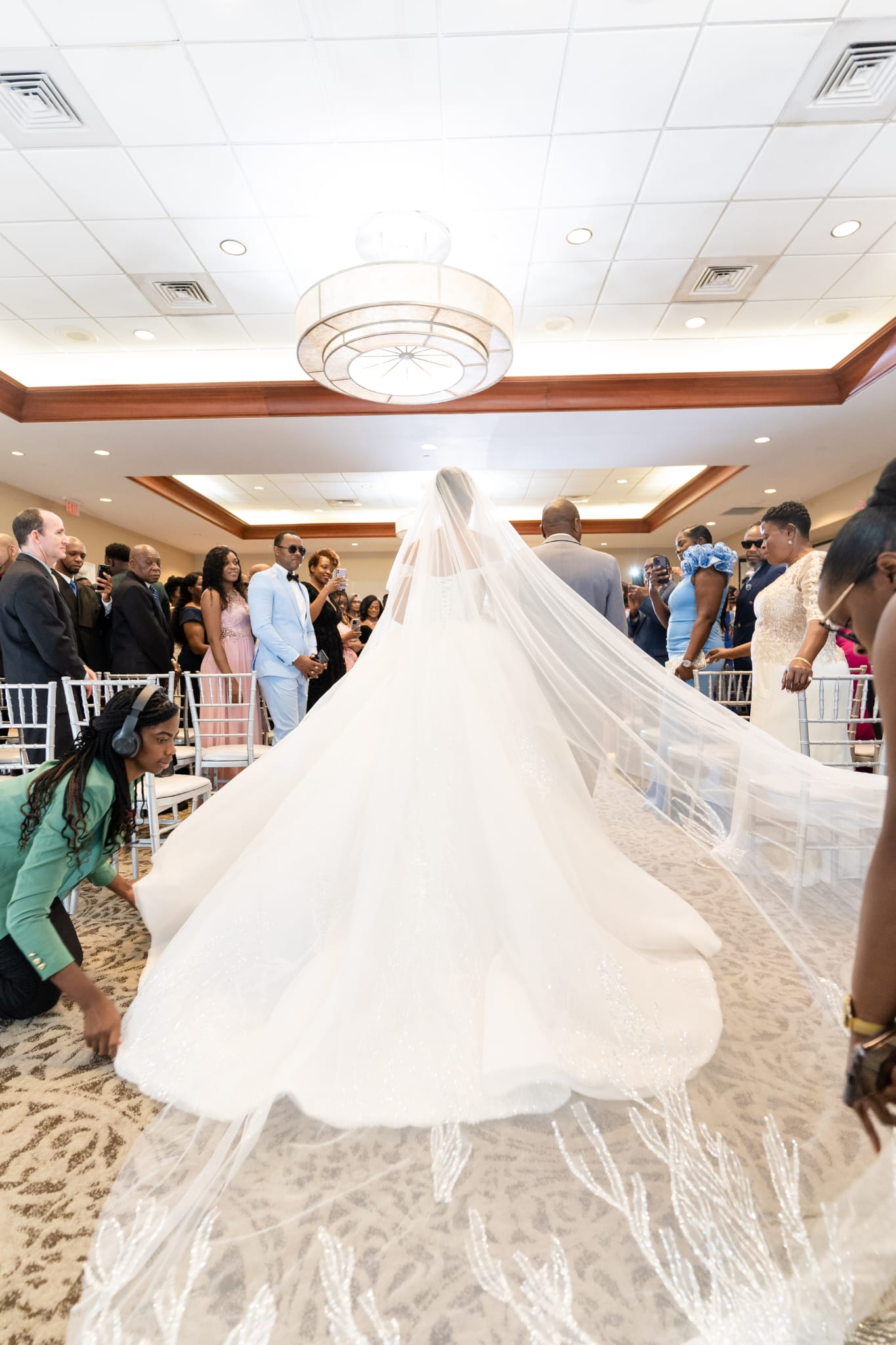 Bride in a wedding gown with a long train, standing in a decorated indoor venue, surrounded by guests.