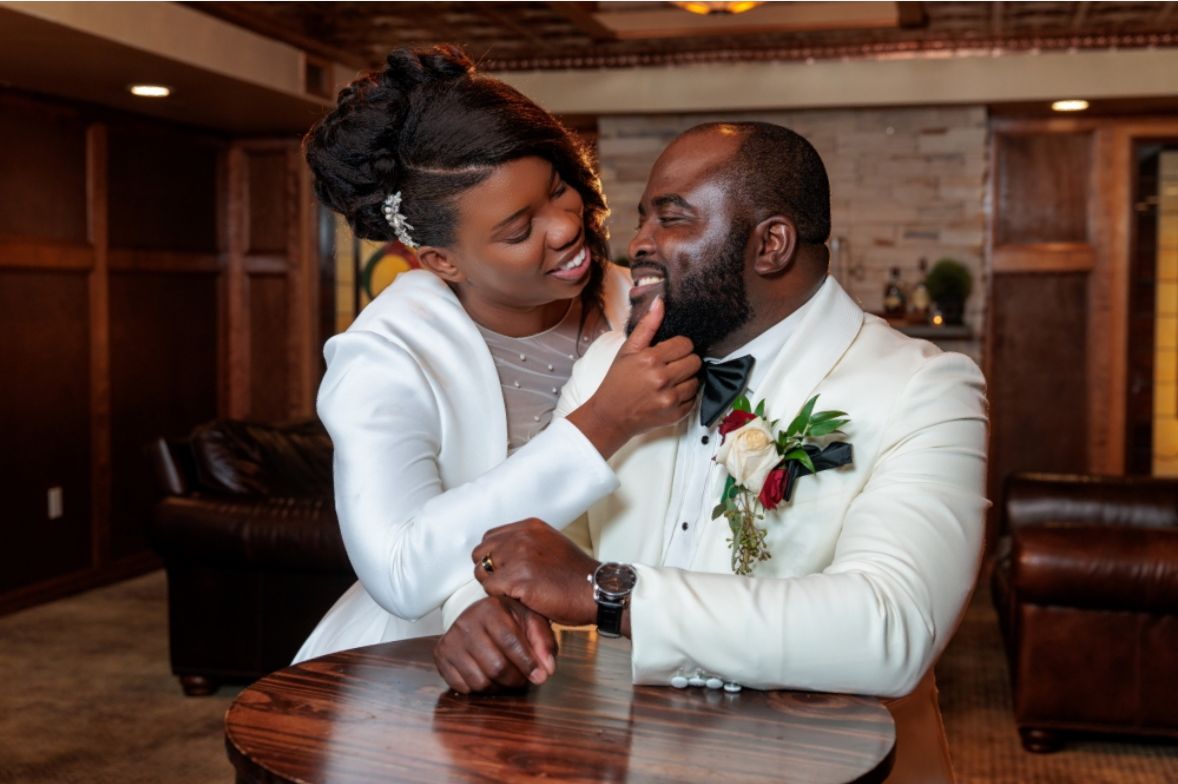 A woman in a white dress touches a man's face, both smiling, in a warmly lit room with wooden furniture.