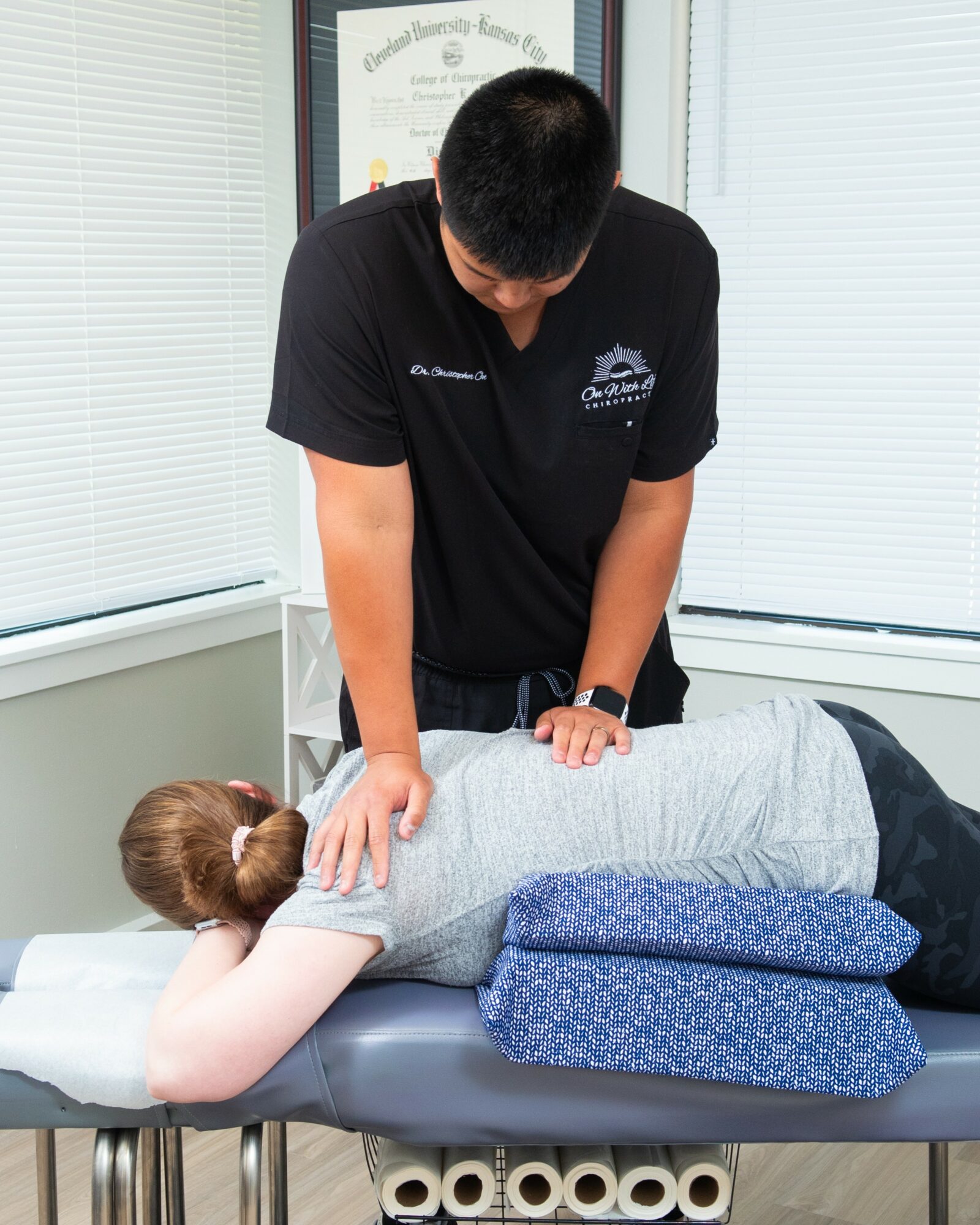 Person receiving a chiropractic adjustment from a practitioner, lying face down on a treatment table with a blue cushion, in a clinical setting.