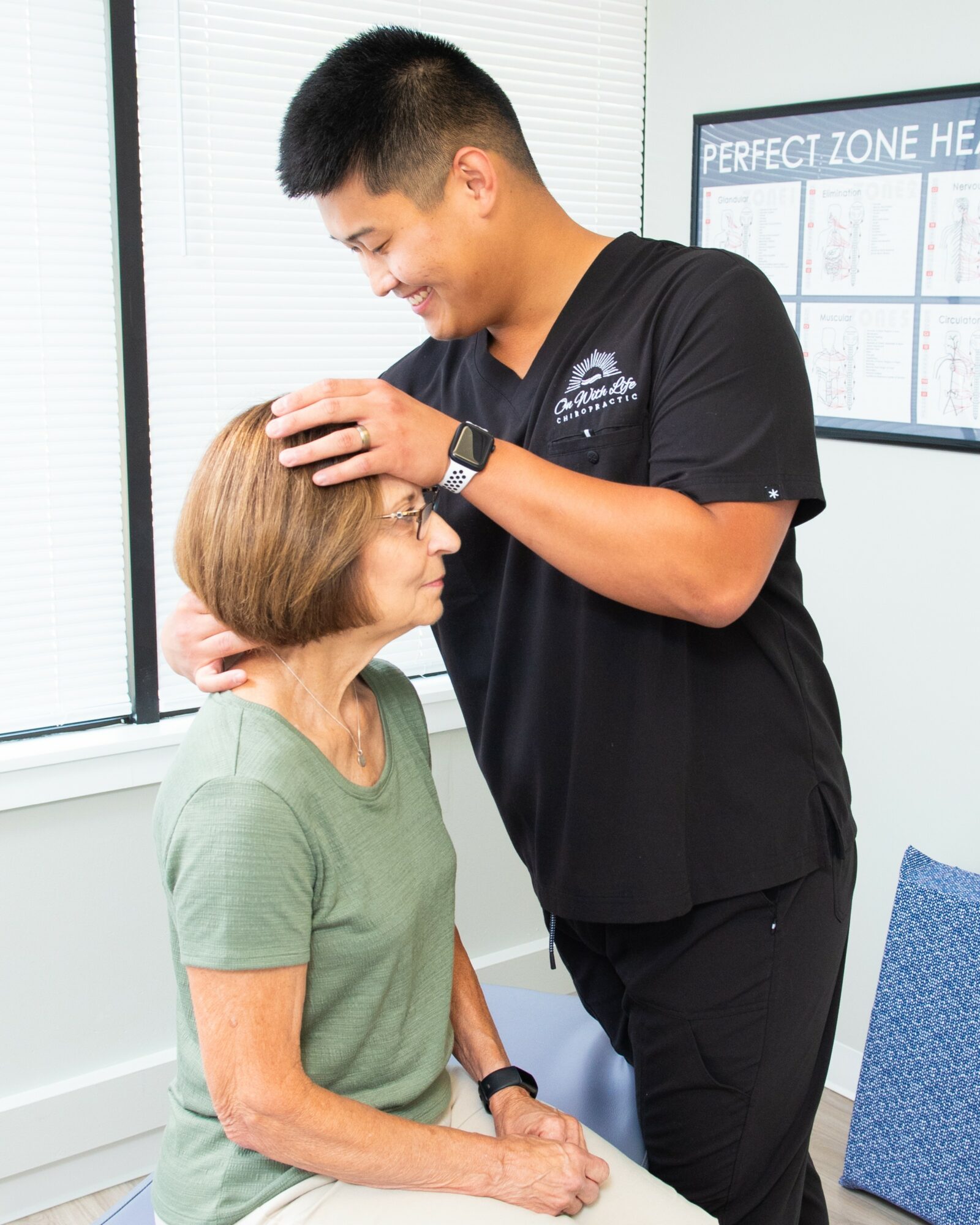 Young man gently touches elderly woman's head in a clinical setting, both smiling, with a window and chart in the background.