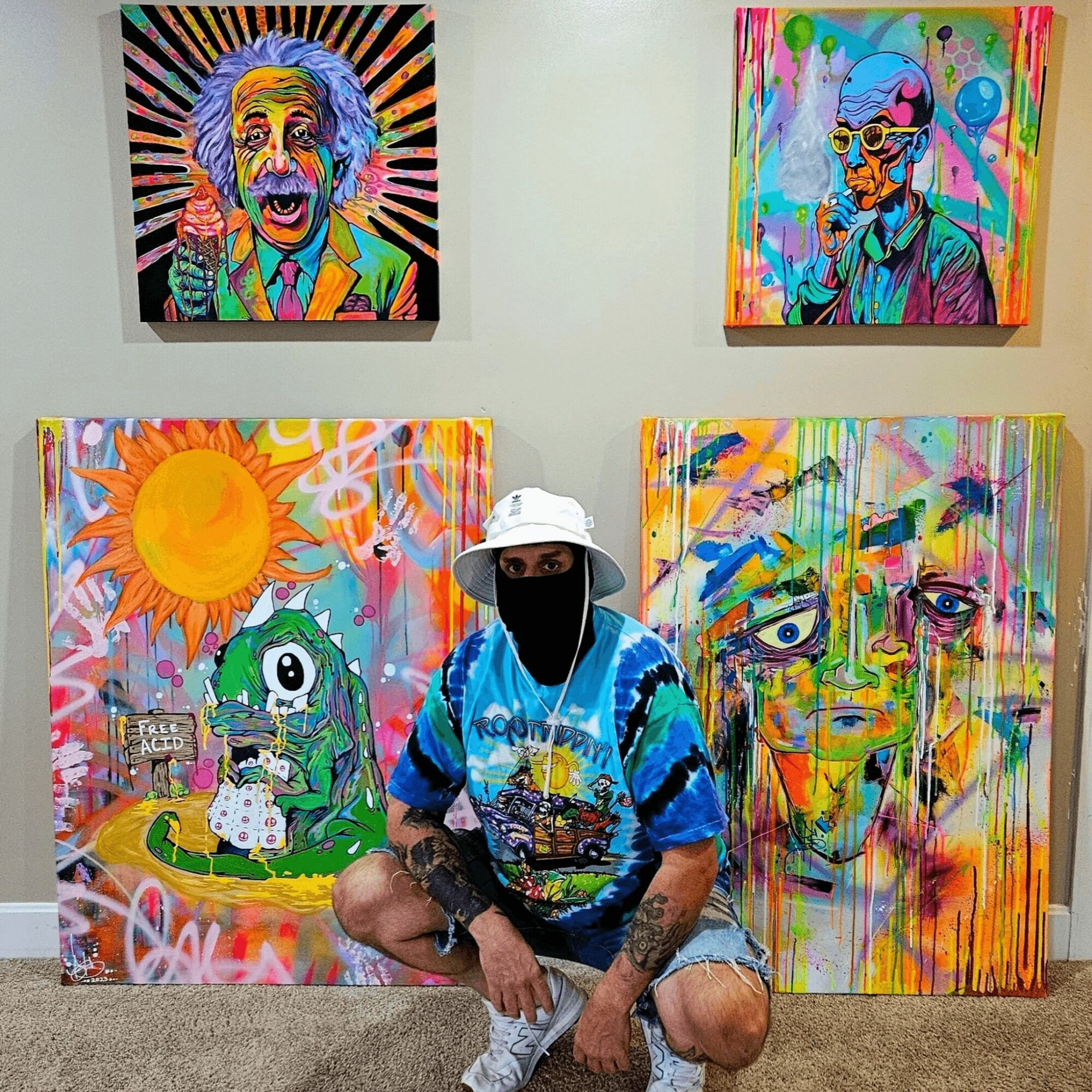 Person sitting in front of colorful abstract paintings, wearing a white hat, blue shirt, and black face mask, with vibrant artwork behind.