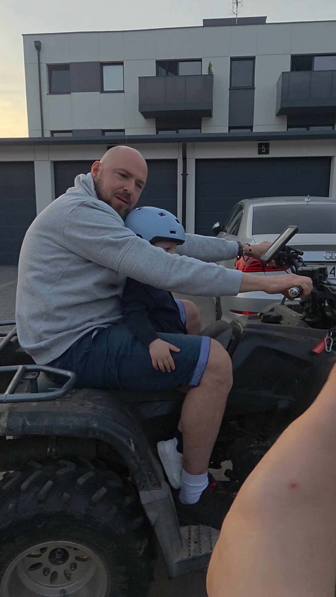 Man sitting on a vehicle, holding a helmet, with a building and parked cars in the background.