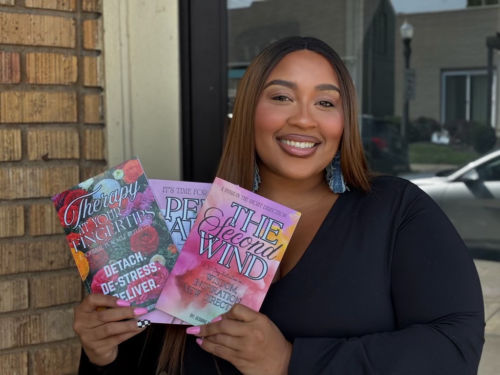 Smiling woman holding three colorful books outside, wearing a black top, with a brick wall and window behind her.