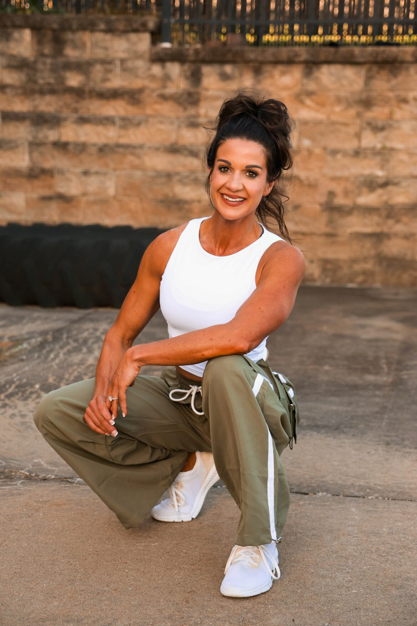Woman crouching outdoors, smiling, wearing a white tank top and khaki pants, with a brick wall background.