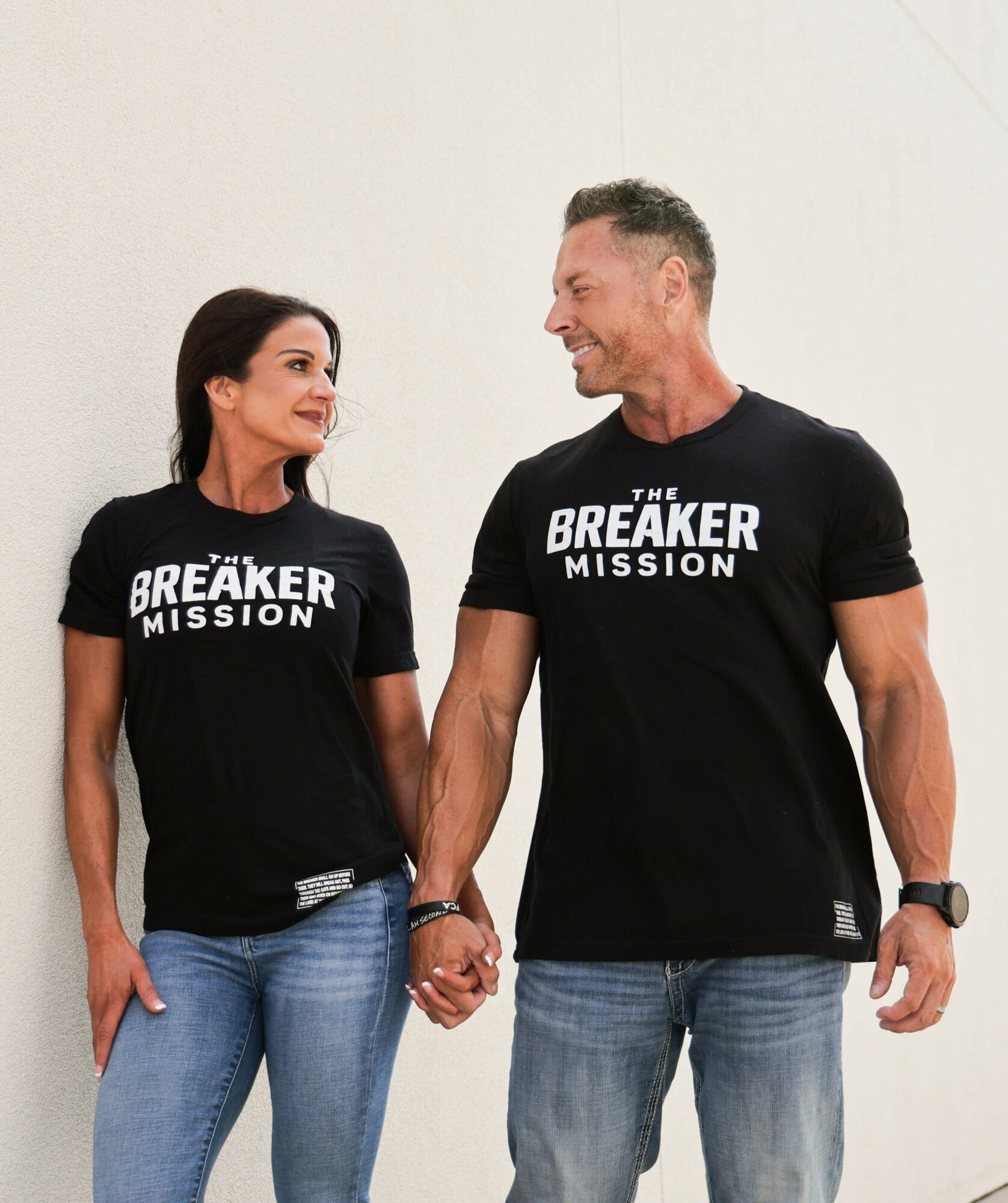 A man and woman hold hands, smiling at each other, wearing matching black T-shirts with white text, standing against a plain white background.