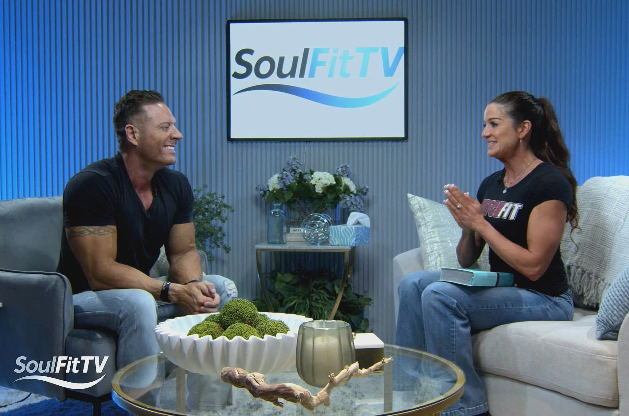 Two people sit on a sofa and talk in a TV studio with a SoulFit TV logo on the wall behind them.