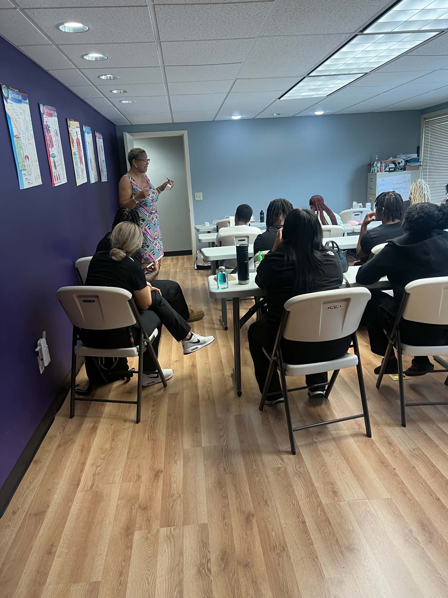 A woman stands at the front of a classroom, speaking to seated adults around tables, with posters on the purple wall.