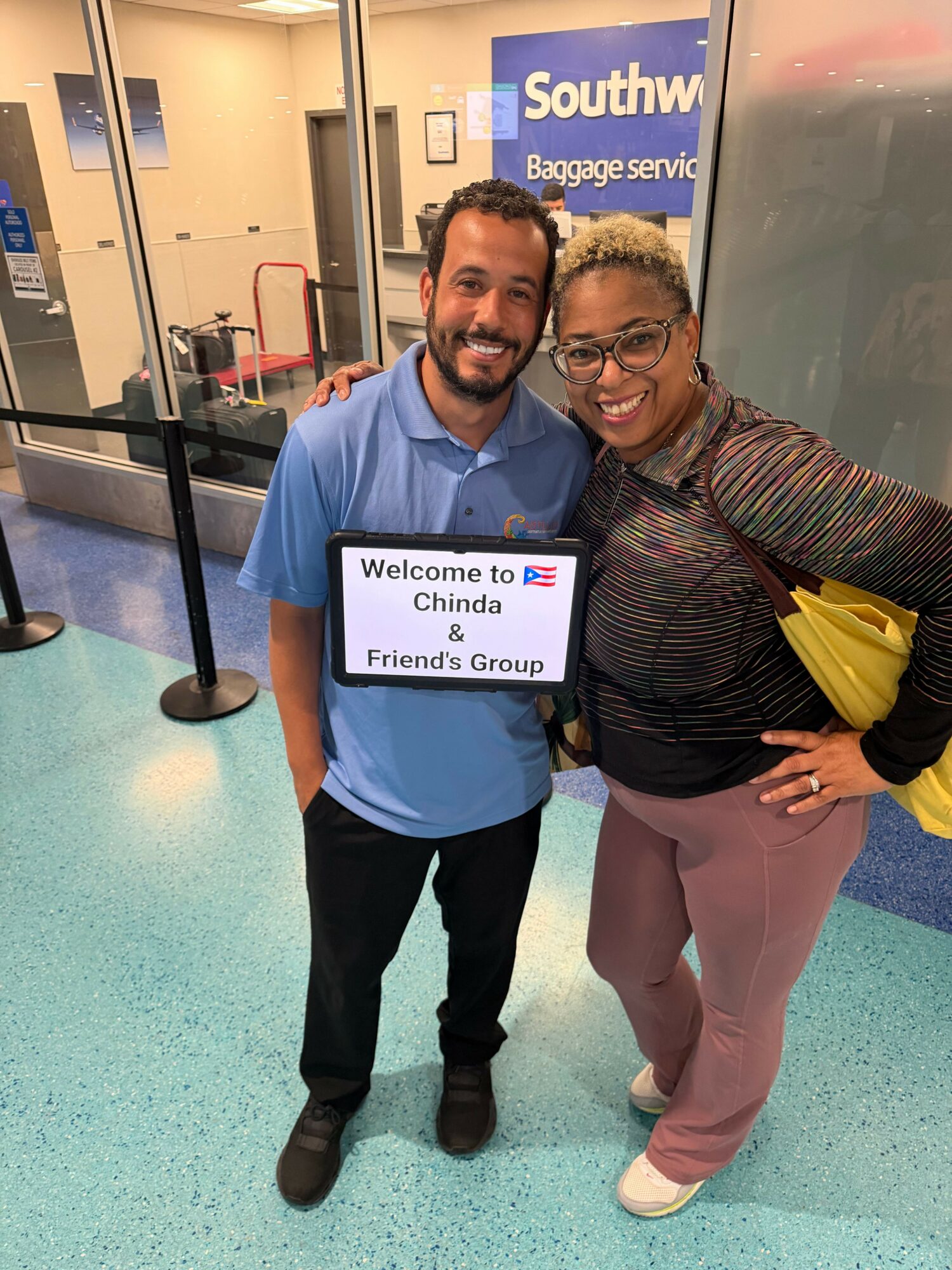 Two people smiling at an airport baggage claim area, holding a welcome sign, with a Southwest Airlines sign in the background.