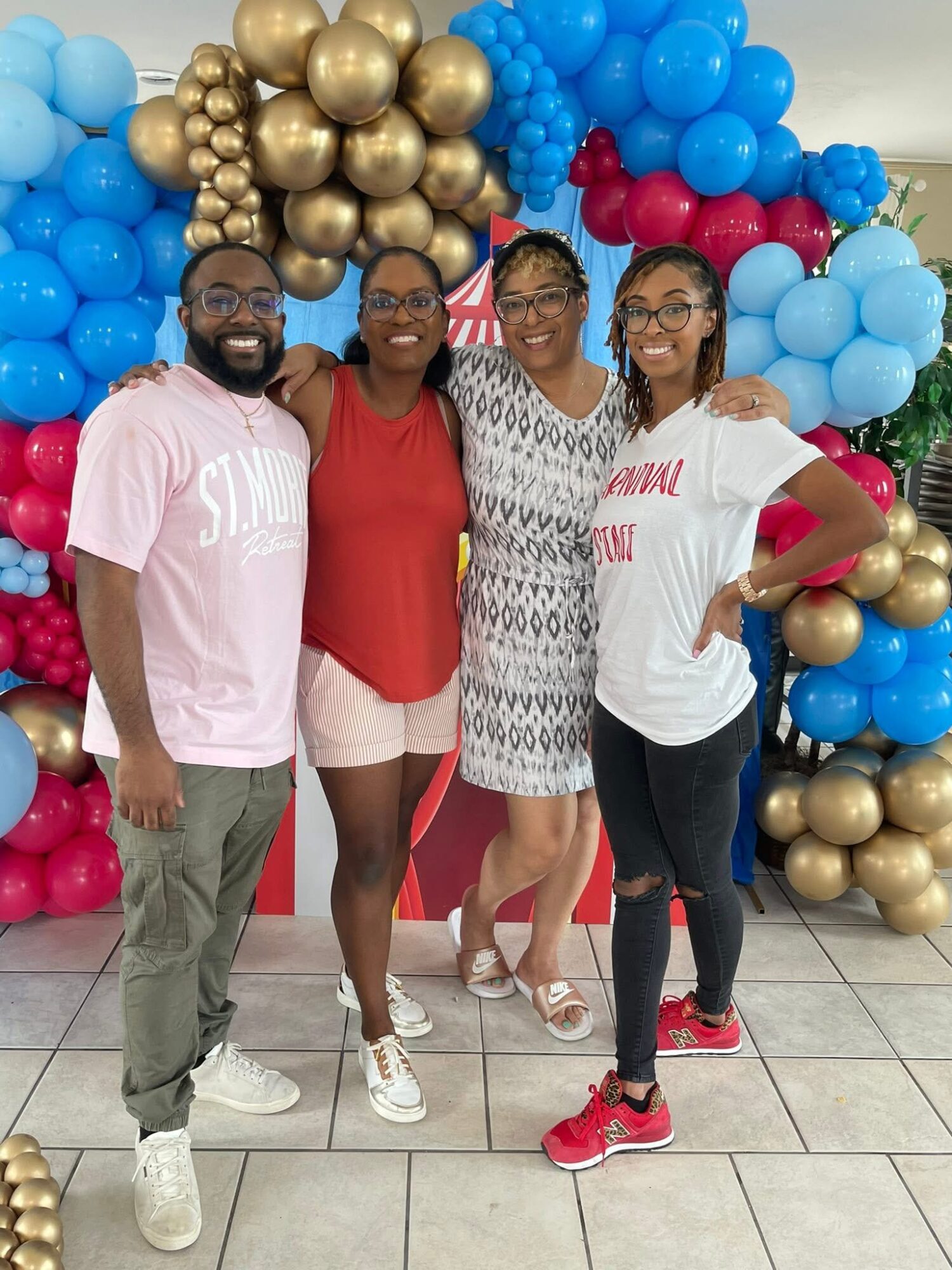 Four people standing in front of a colorful balloon arch, smiling, with a tiled floor, celebrating an event.