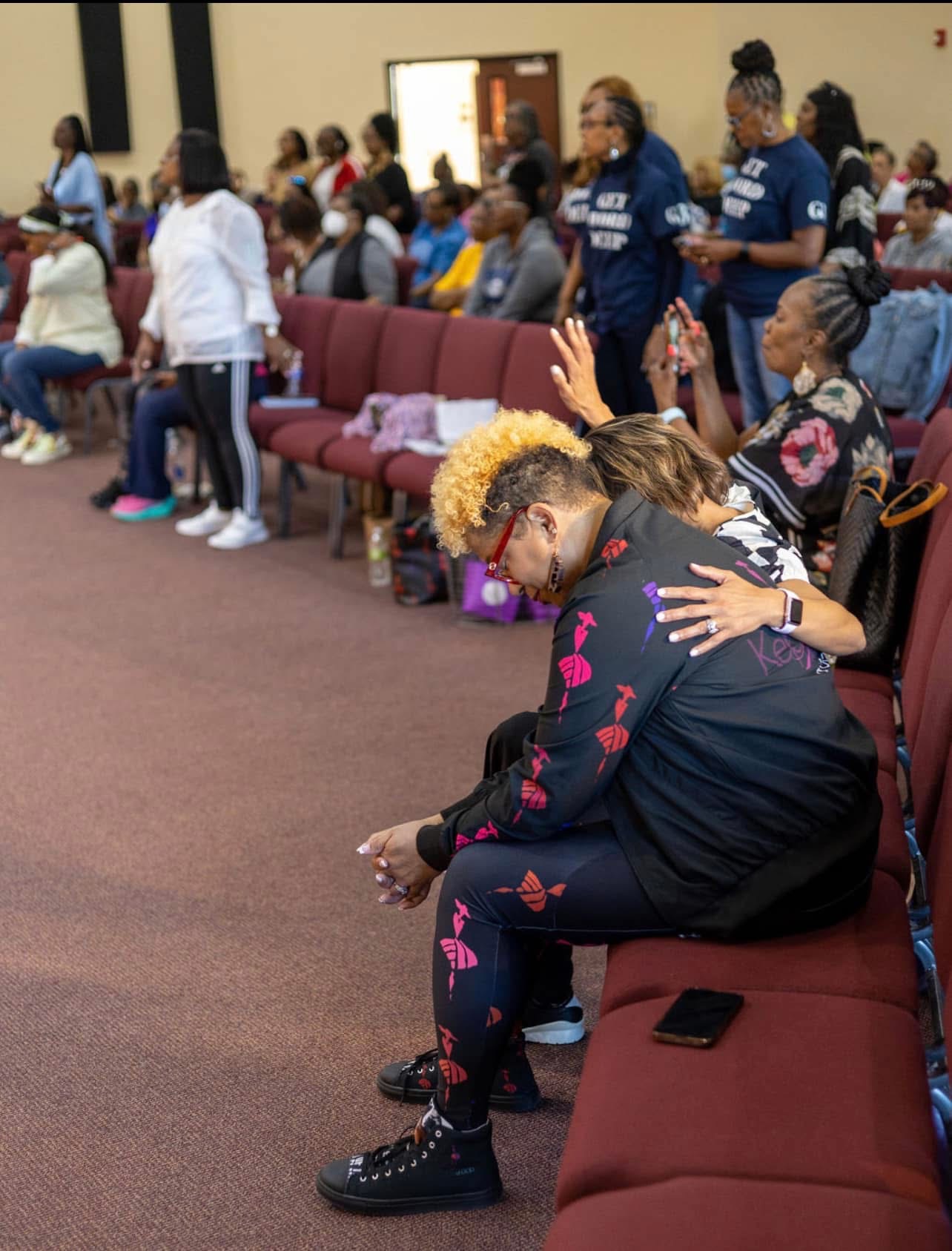 Two women sit on a bench, one with a yellow hairstyle and the other with braided hair, in a crowded room with people standing and sitting. 20 words.