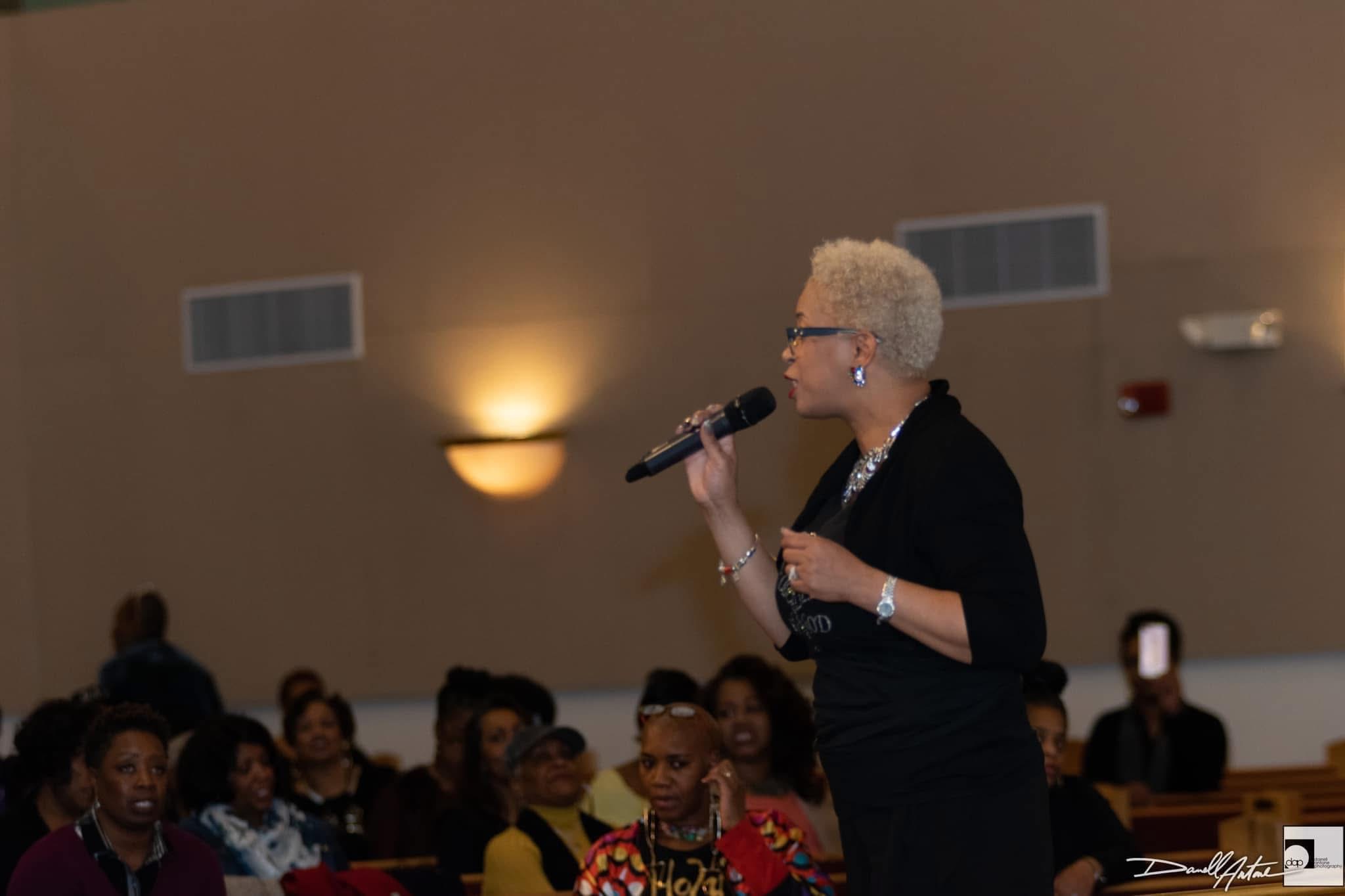 Woman with short blonde hair, glasses, black outfit, speaking into a microphone in a room with seated audience.