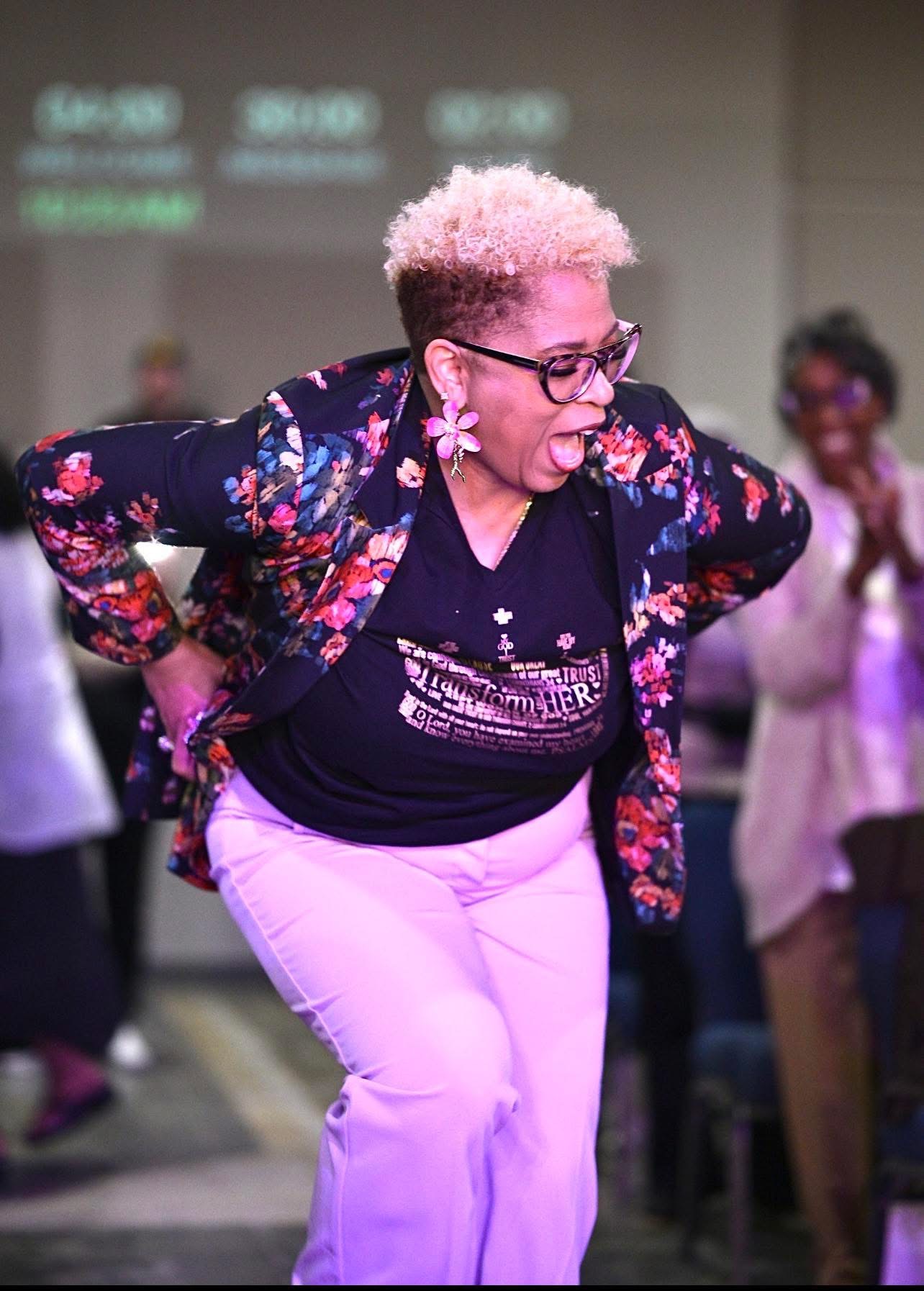 Person with short, curly pink hair and glasses laughing, wearing a floral jacket and light-colored pants, in a group setting.