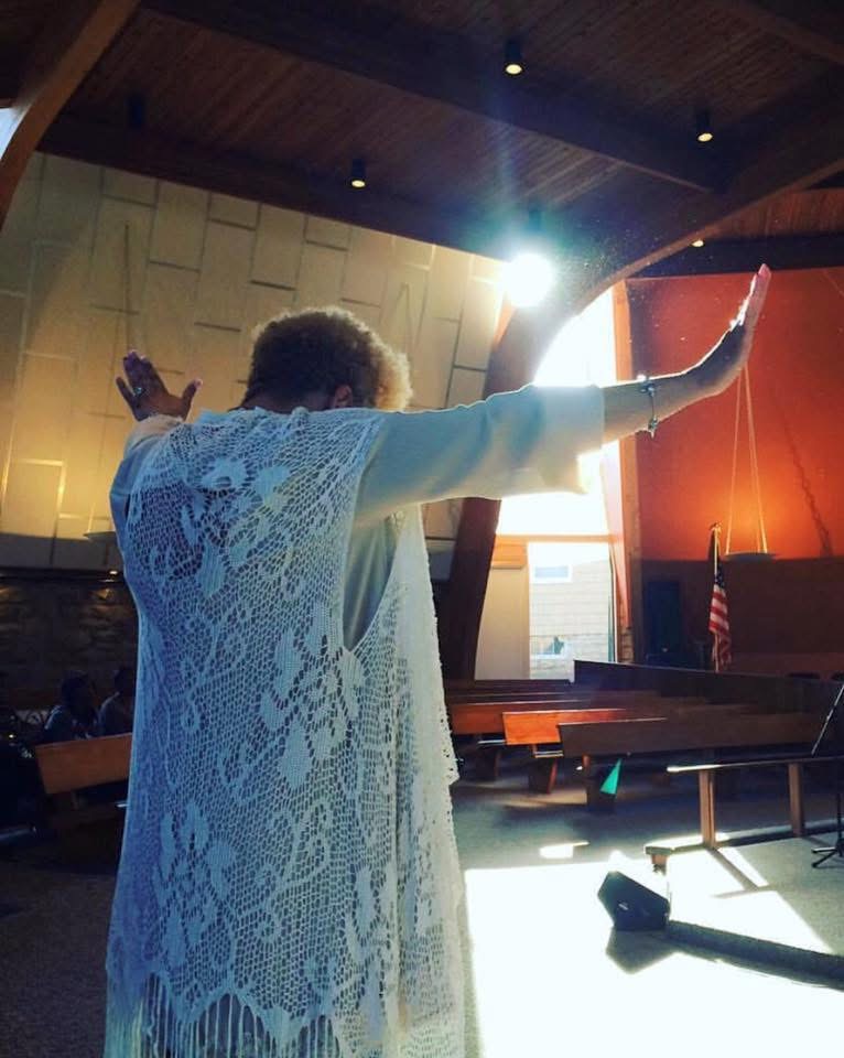 Person with arms raised, wearing a lace garment, standing in a room with wooden ceiling and bright light behind.