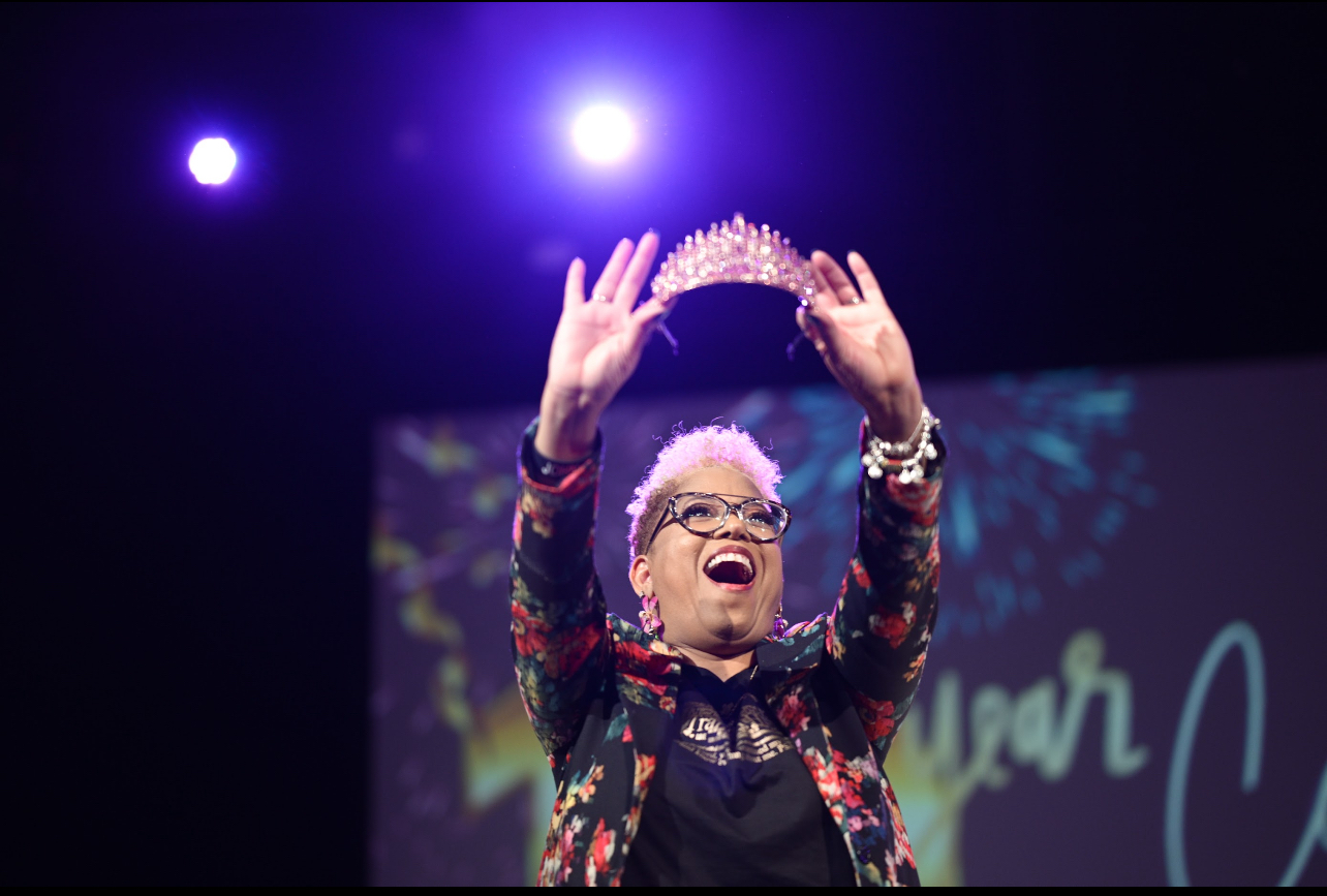 Woman wearing glasses and a crown, smiling and raising her hands on stage with purple lights behind her.