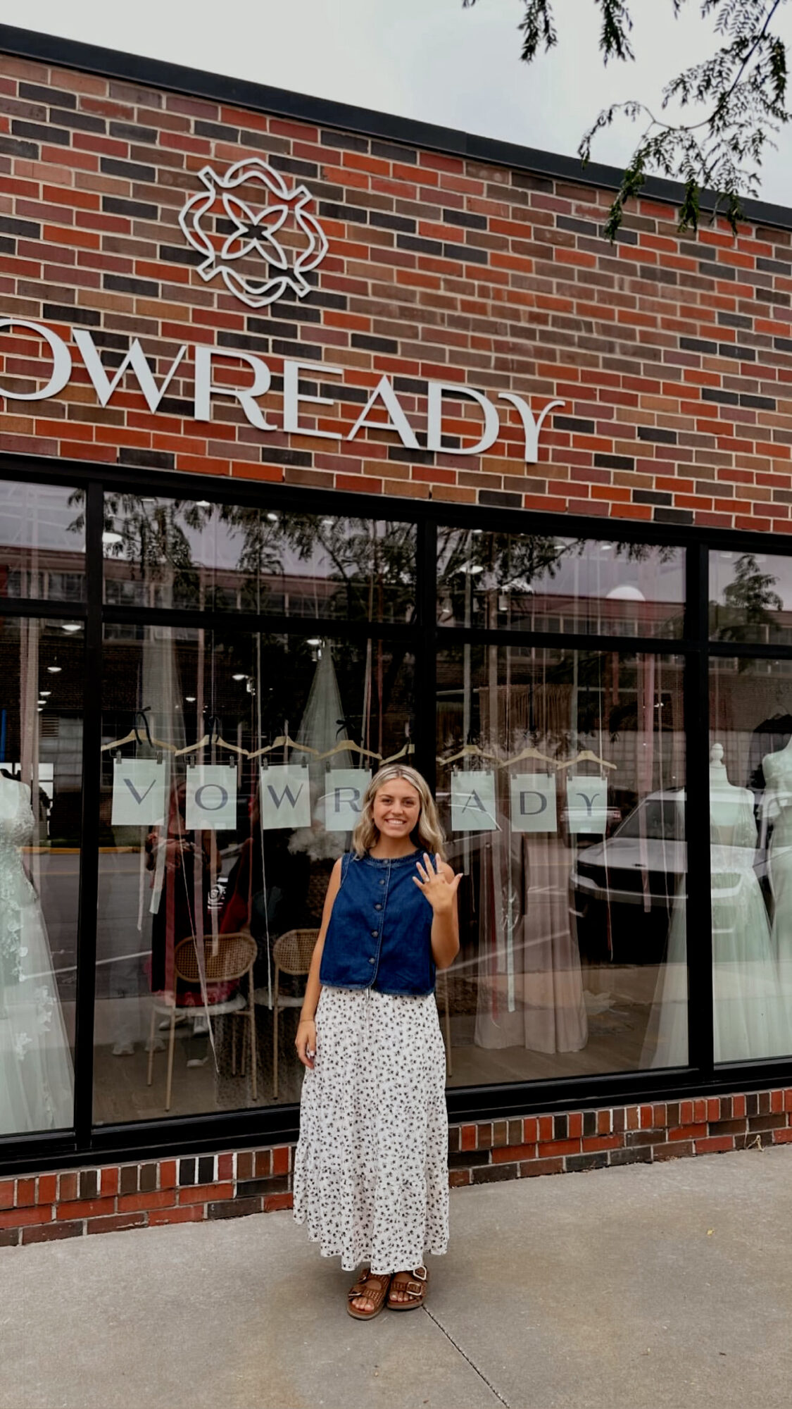Woman standing outside a brick building with large glass windows and a sign reading 'OWRE ADY'.