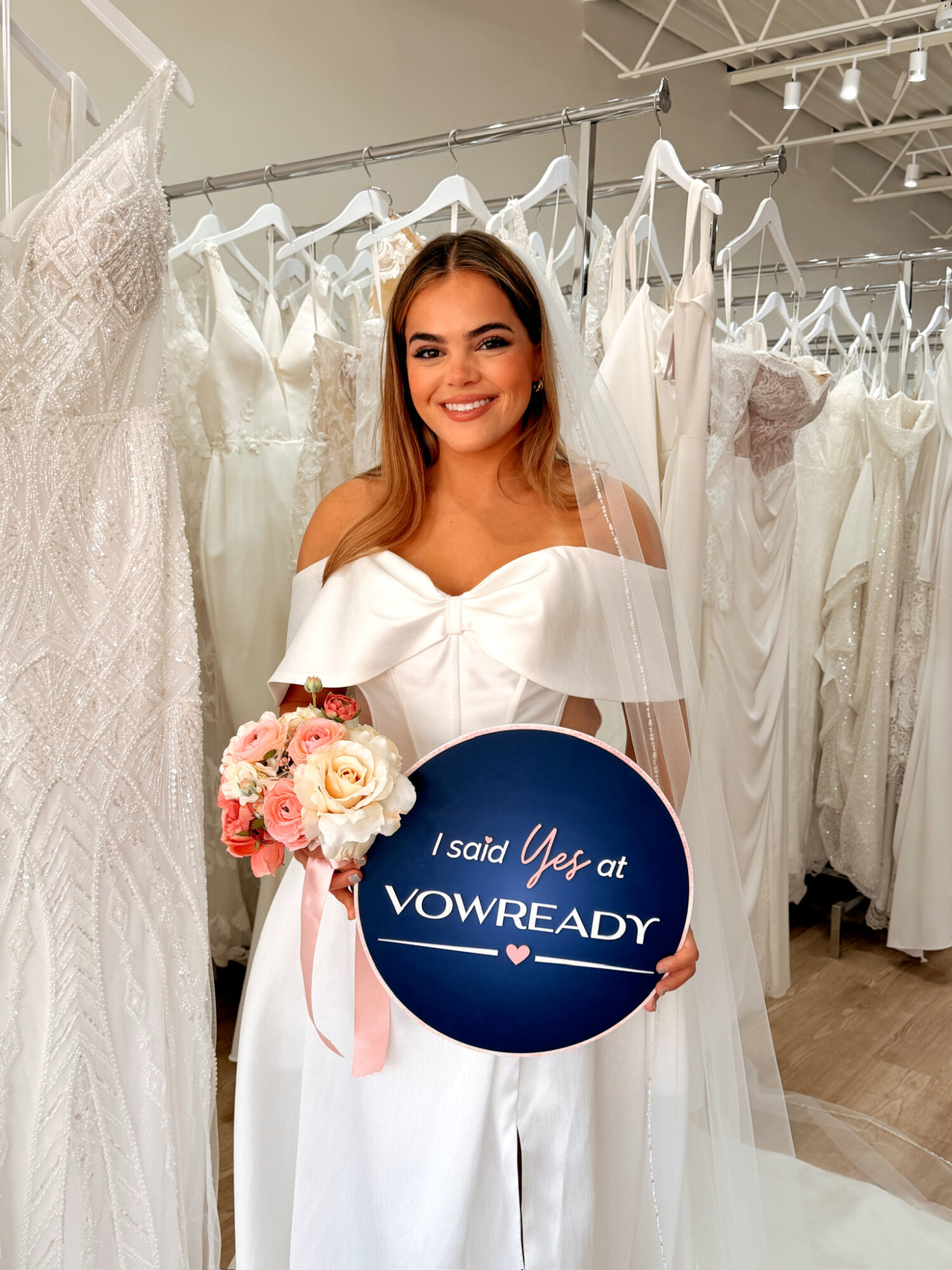 Young woman in a white dress holding a bouquet and a sign in a bridal shop, surrounded by wedding dresses.