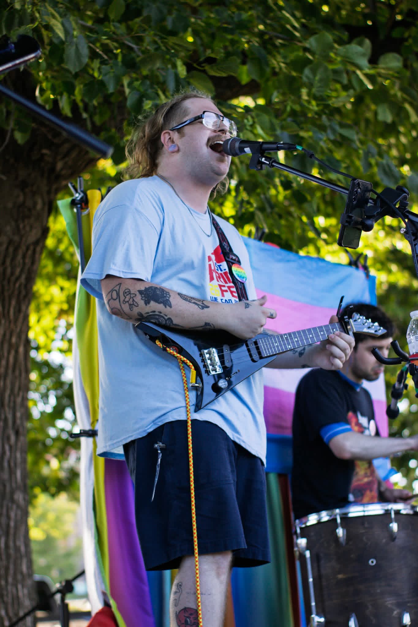 Person with glasses singing into a microphone while playing an electric guitar outdoors, with a person playing drums in the background.