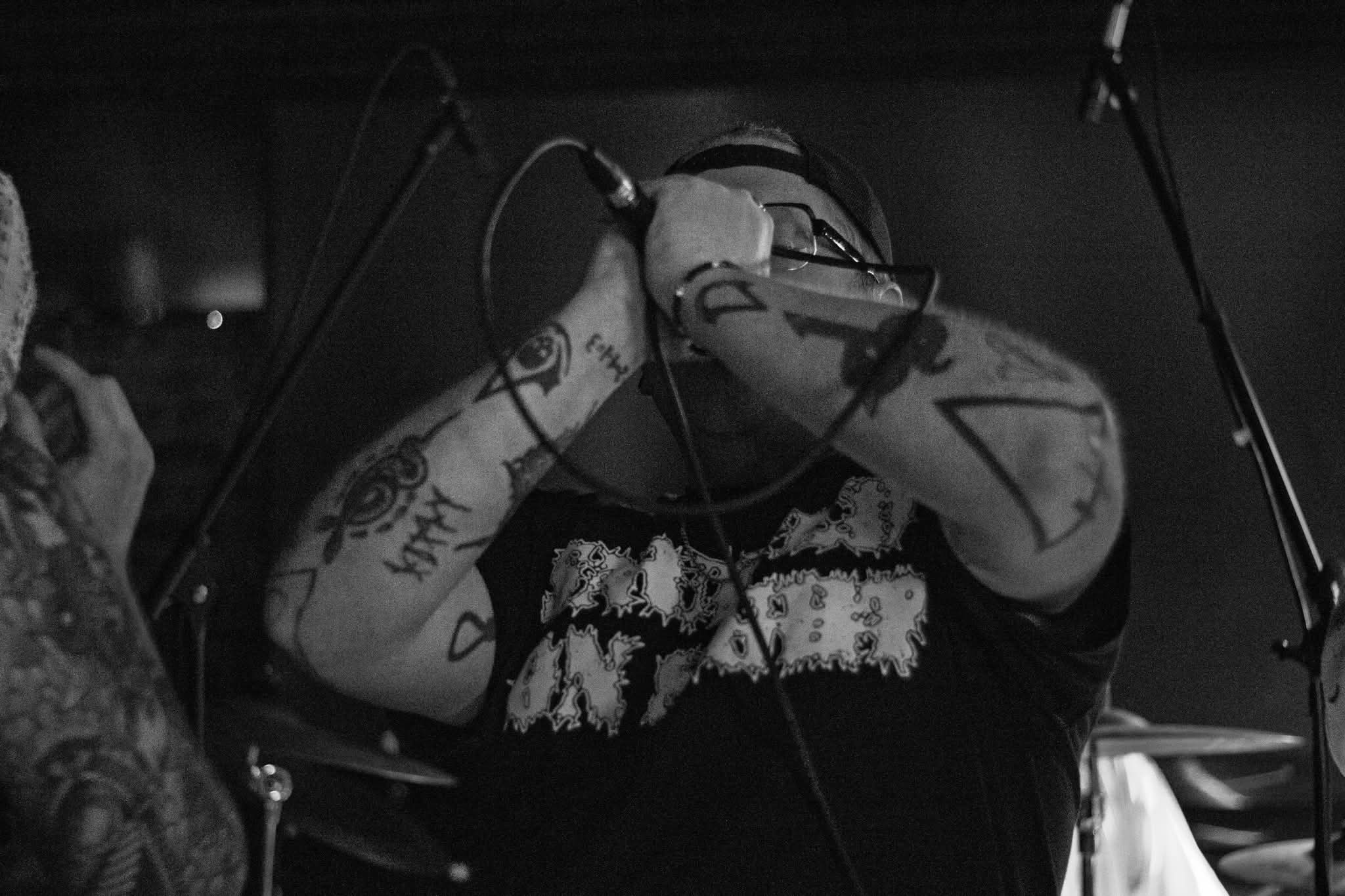 Person with tattoos covering arms, holding head with hands, in a dark setting, possibly a music studio or stage.