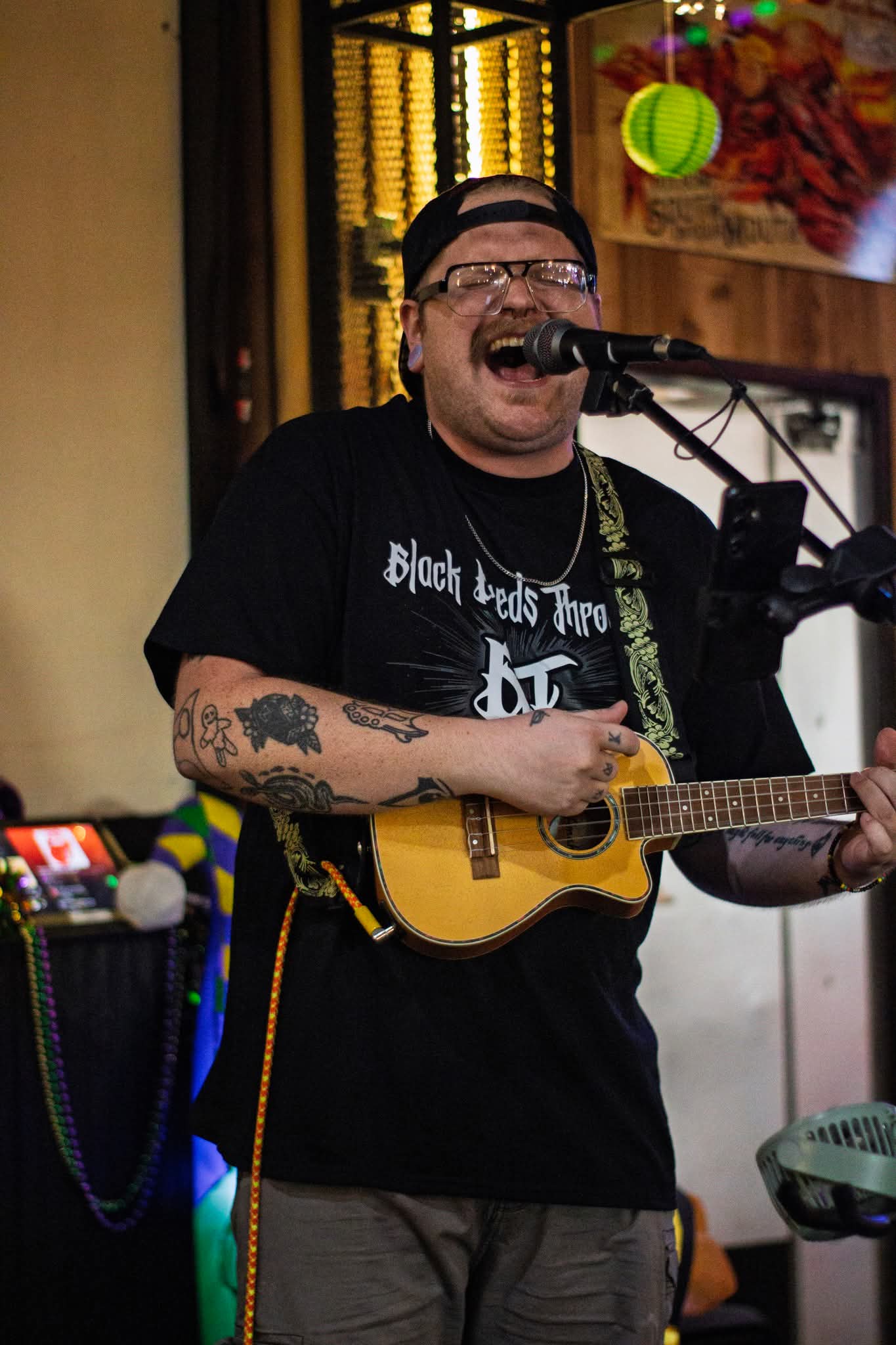 Person singing and playing guitar with microphone, wearing glasses and a black T-shirt, tattoos on arms, in indoor setting.