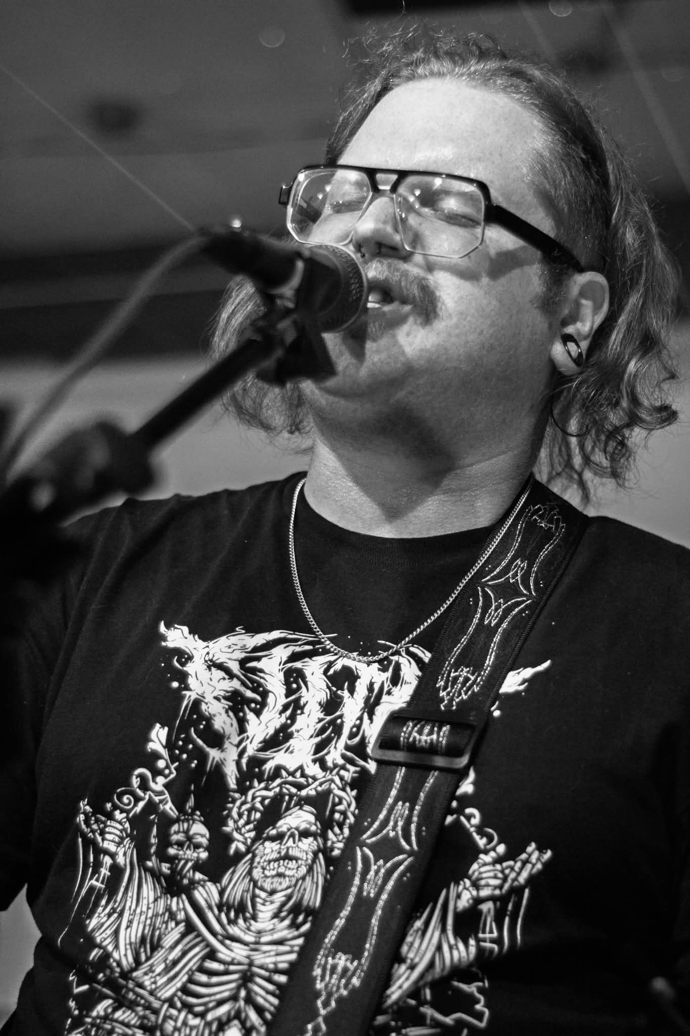 Person singing into microphone, wearing glasses and a graphic t-shirt, with a guitar strap visible.