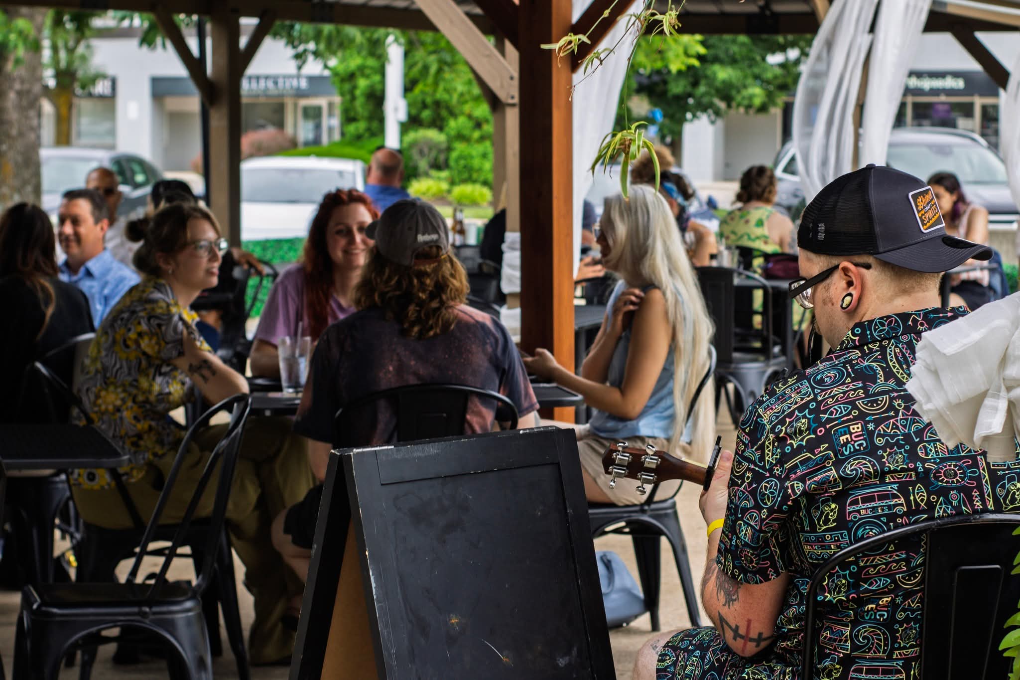 Group of people sitting at outdoor tables under a wooden structure, engaged in conversation, with trees and buildings in background.