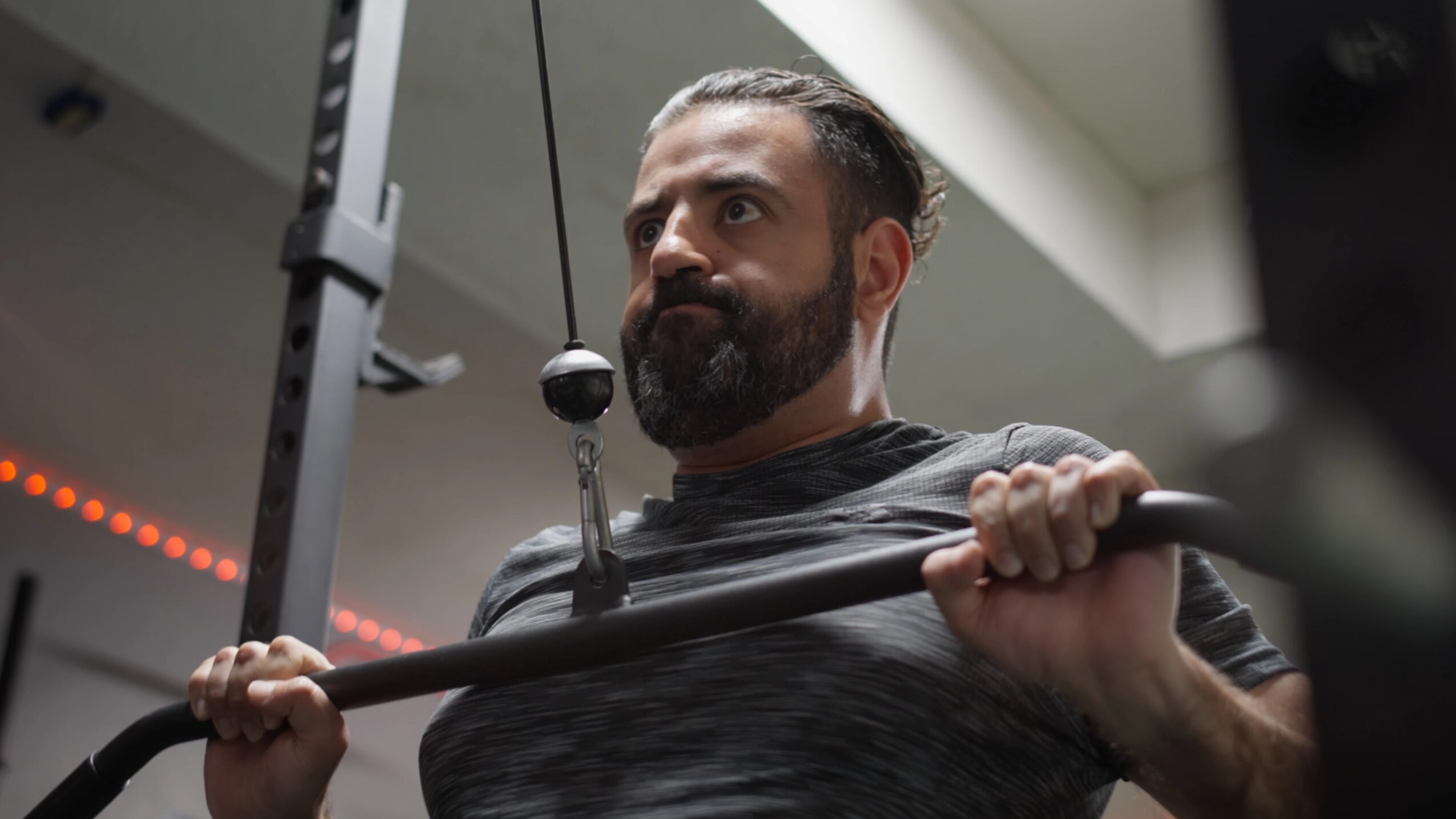 Man with a beard lifting a barbell in a gym, focused expression, indoor setting with ceiling light.